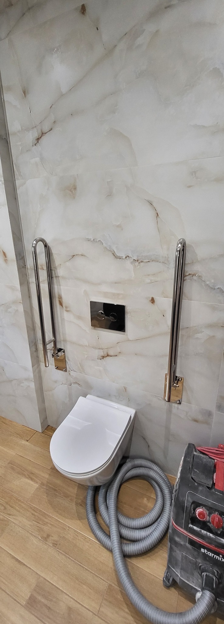 Nowoczesna toaleta podwieszana z chromowanymi uchwytami dla niepełnosprawnych na marmurowej ścianie. Na podłodze widoczny odkurzacz przemysłowy.