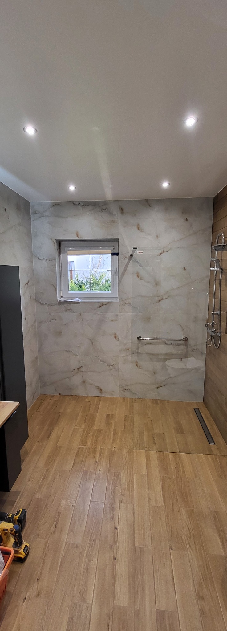 Wykończona łazienka z marmurowymi płytkami ściennymi i drewnopodobną podłogą. Widoczne okno, armatura prysznicowa i narzędzia glazurnicze w rogu.