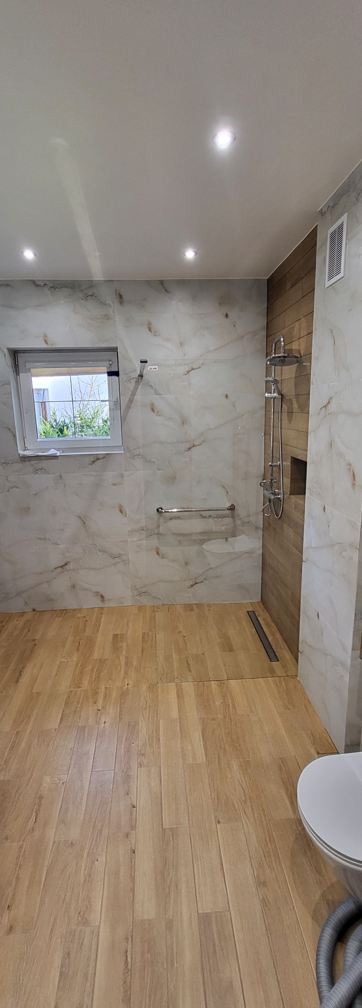 Wykończona łazienka z drewnopodobną podłogą i marmurowymi płytkami ściennymi. Kabina prysznicowa typu walk-in z odpływem liniowym. Widoczne okno i chromowana armatura.
