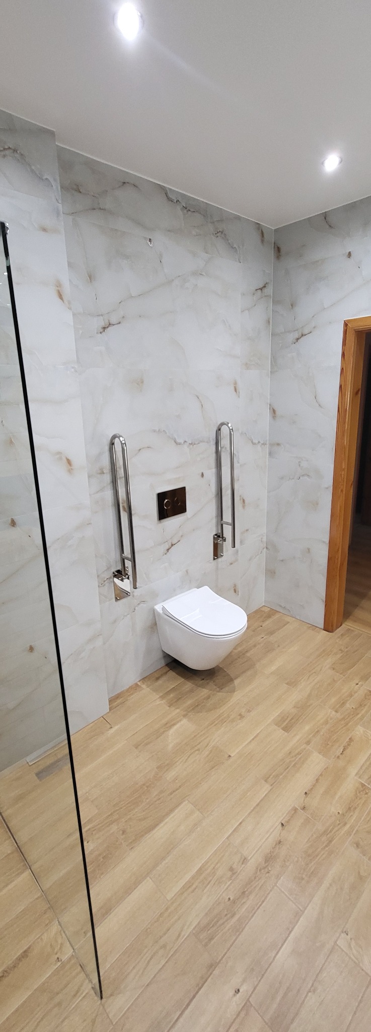 Jasna łazienka z marmurowymi płytkami, drewnopodobną podłogą, toaletą i uchwytami dla niepełnosprawnych. Minimalistyczny design, nowoczesne oświetlenie.
