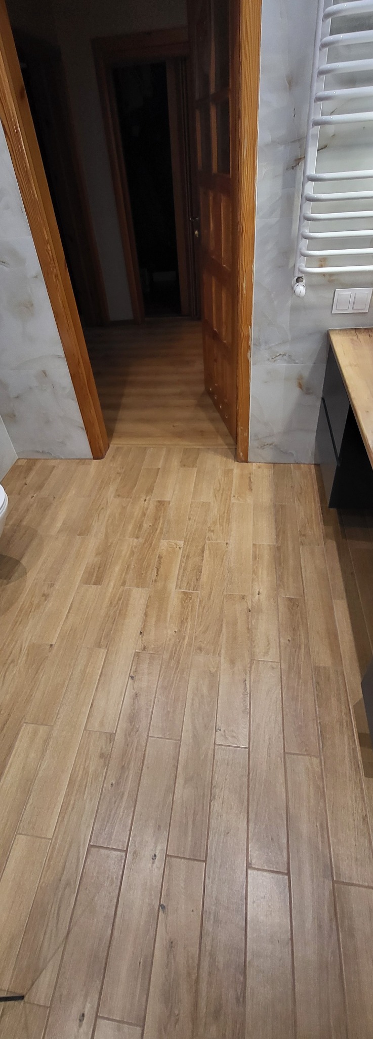 Ułożone płytki podłogowe imitujące drewno w łazience, widok z góry, z fragmentem grzejnika i otwartymi drzwiami w tle. Jasne, ciepłe kolory.