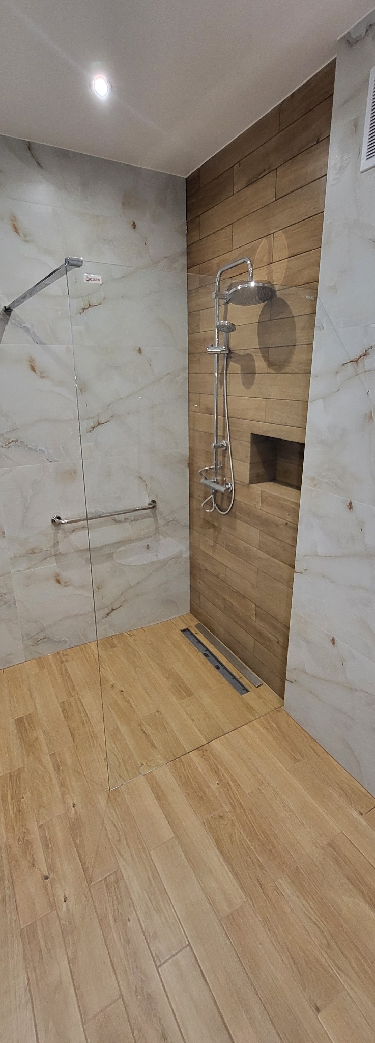 Nowoczesna łazienka z kabiną typu walk-in, wykończona płytkami imitującymi drewno i marmur. Widoczna armatura prysznicowa i odpływ liniowy.
