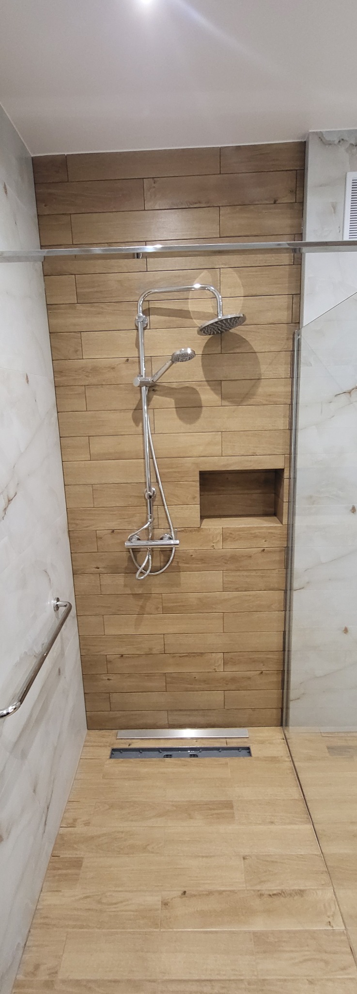 Nowoczesna kabina prysznicowa z drewnopodobnymi płytkami, chromowaną armaturą i odpływem liniowym. W ścianie wnęka na kosmetyki. Jasne, marmurowe płytki po bokach.