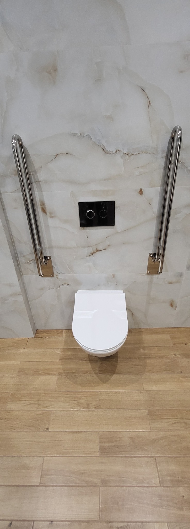 Wykończona łazienka z podwieszaną toaletą, chromowanymi uchwytami dla niepełnosprawnych i marmurowymi płytkami ściennymi. Ujęcie z góry.