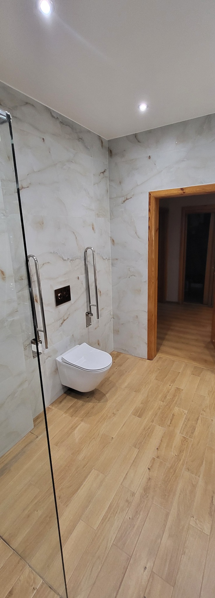 Wykończona łazienka z beżowymi płytkami imitującymi drewno na podłodze i ścianach. Wisząca toaleta z uchwytami dla niepełnosprawnych. Kabina prysznicowa ze szklaną ścianką.