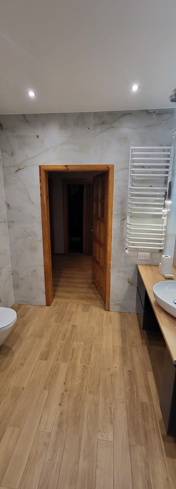 Wykończona łazienka z drewnopodobną podłogą, marmurowymi płytkami ściennymi i drewnianą ościeżnicą drzwiową. Grzejnik drabinkowy i umywalka na blacie.