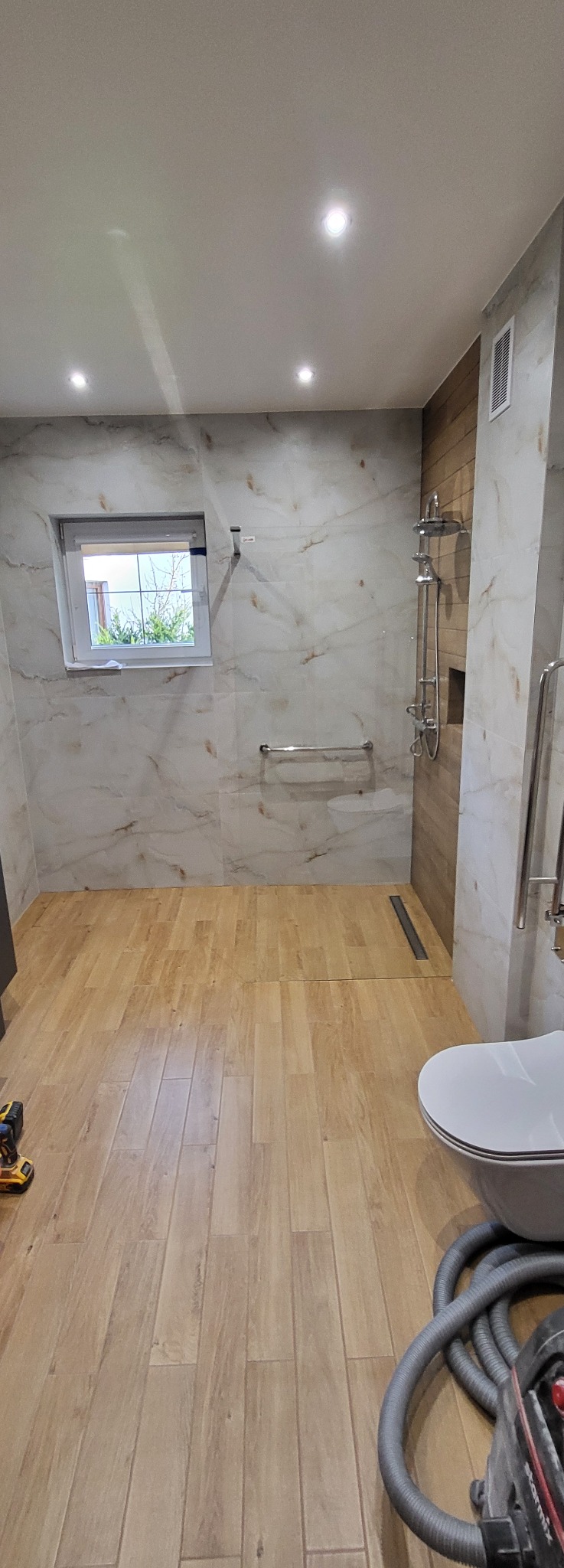 Wykończona łazienka z drewnopodobną podłogą, marmurowymi płytkami ściennymi i kabiną prysznicową typu walk-in. Widoczne narzędzia sugerują trwające prace wykończeniowe.