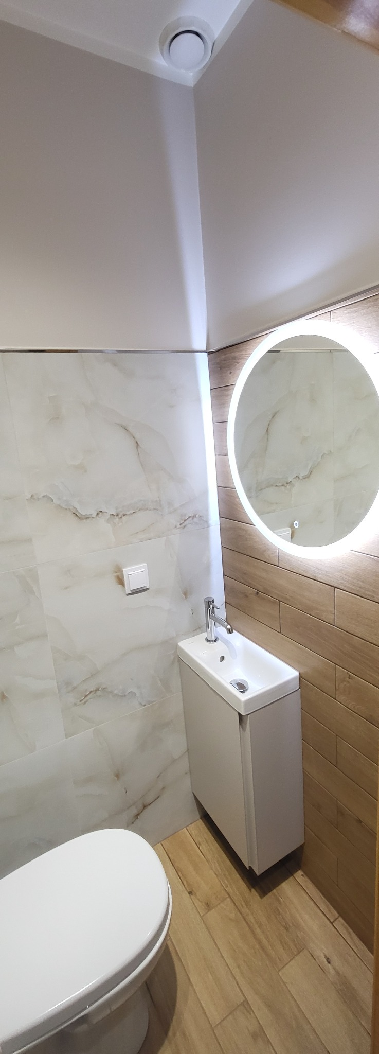 Wykończona łazienka z umywalką na szafce, podświetlanym lustrem i marmurowymi płytkami. Widoczny fragment toalety i drewnopodobna podłoga.