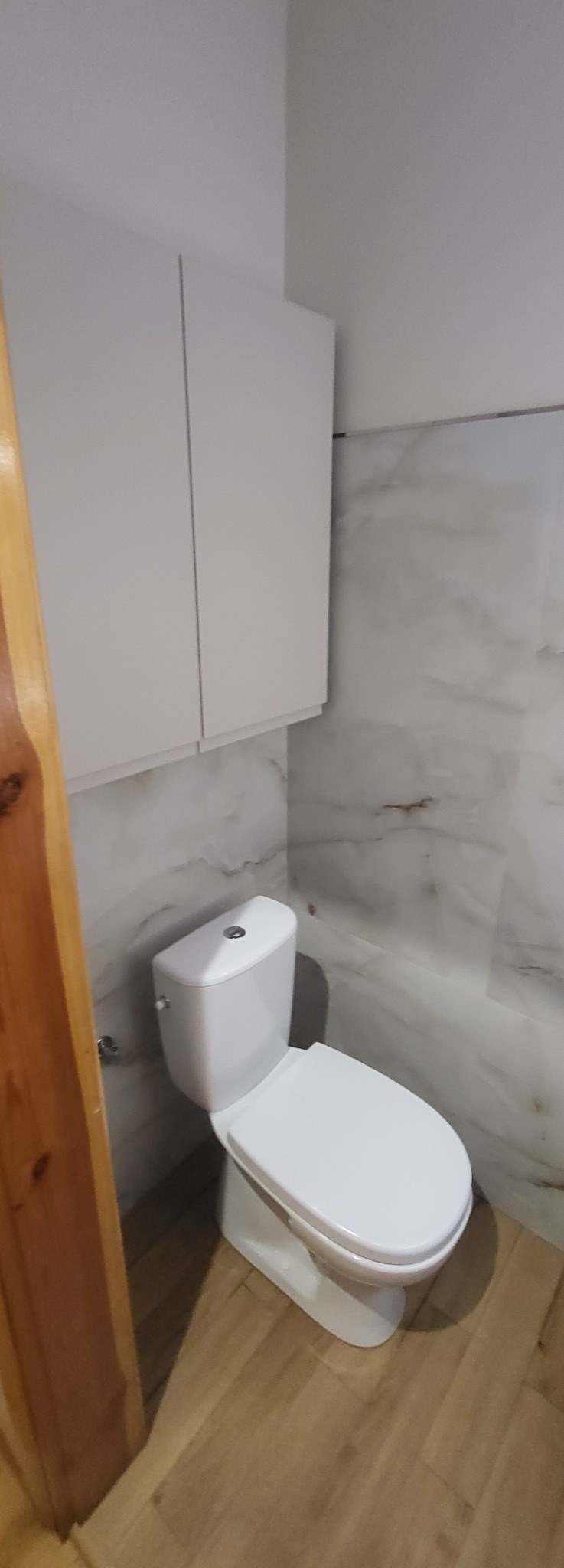 Biała toaleta kompaktowa z szafką wiszącą w wykończonej łazience. Marmurowe płytki ścienne i podłoga imitująca drewno. Widoczny fragment drewnianej futryny.