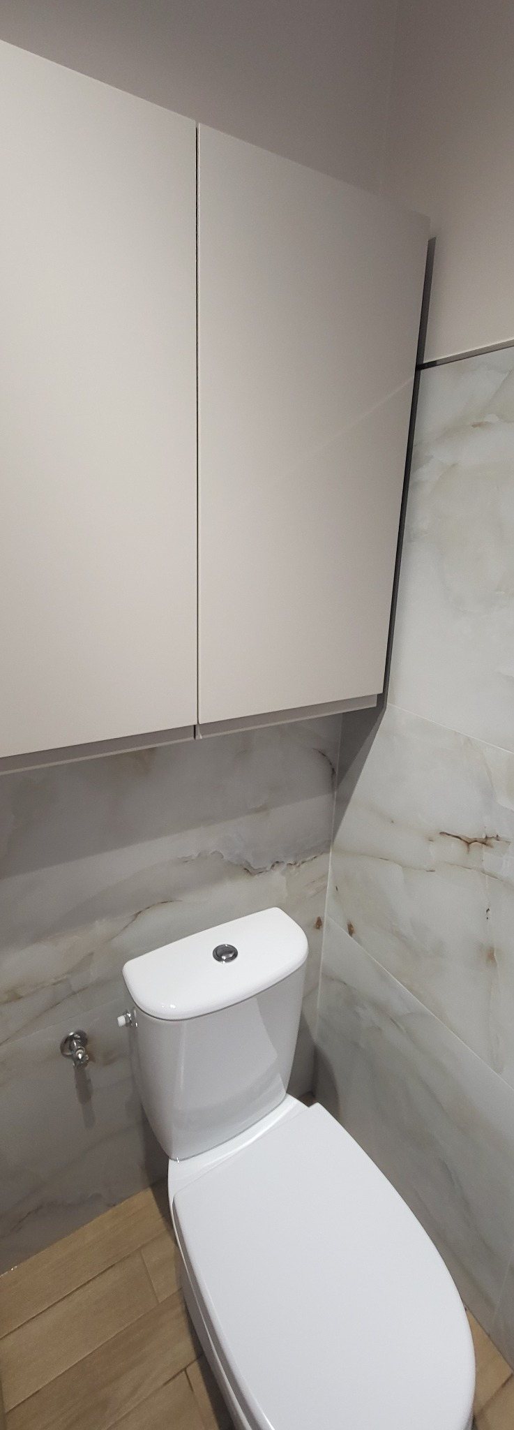 Biała toaleta kompaktowa z szafką wiszącą w wykończonej łazience. Marmurowe płytki na ścianach i drewnopodobna podłoga. Minimalistyczny design.