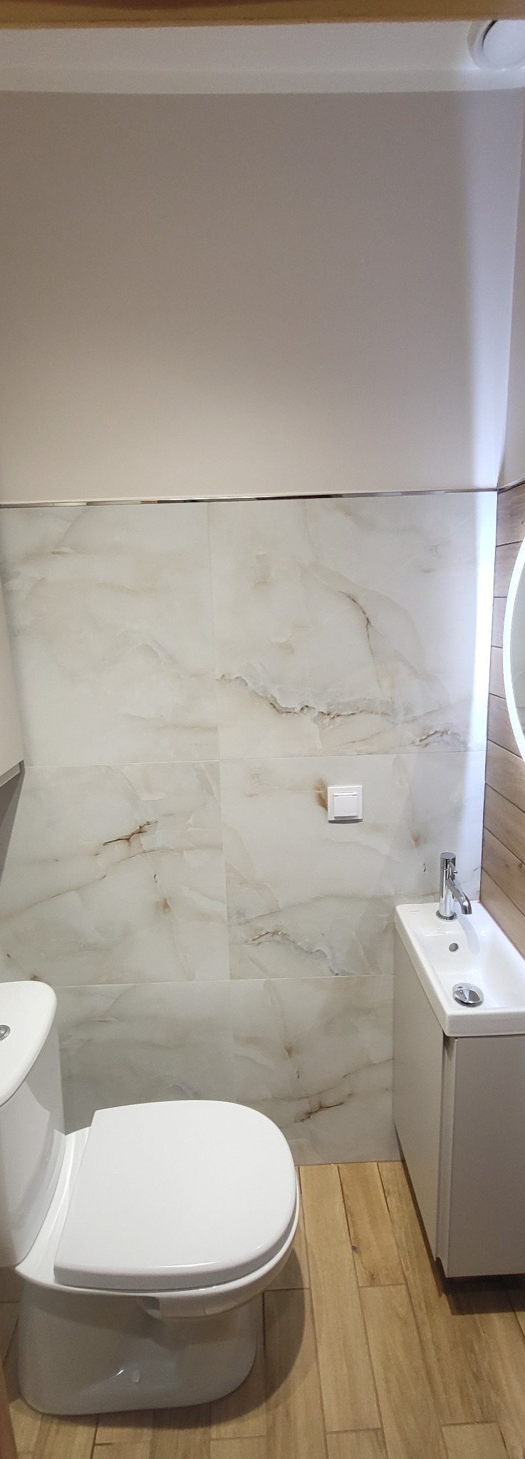 Wykończona łazienka z marmurowymi płytkami na ścianach, drewnopodobną podłogą, białą toaletą i umywalką z szafką. Widoczny fragment lustra z podświetleniem.