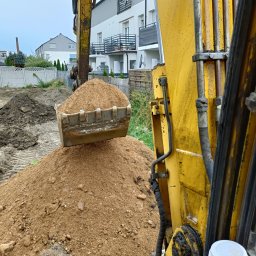 P.D DROBUD Dawid Marszałek - Żółta koparka z pełną łyżką piasku, widok z kabiny, w tle domy jednorodzinne i ogrodzenie.