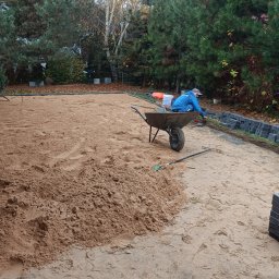 P.D DROBUD Dawid Marszałek - Przygotowanie podłoża pod nawierzchnię: wyrównany piasek, częściowo ułożone obrzeża z kostki, taczka i grabie w ogrodzie z drzewami iglastymi.