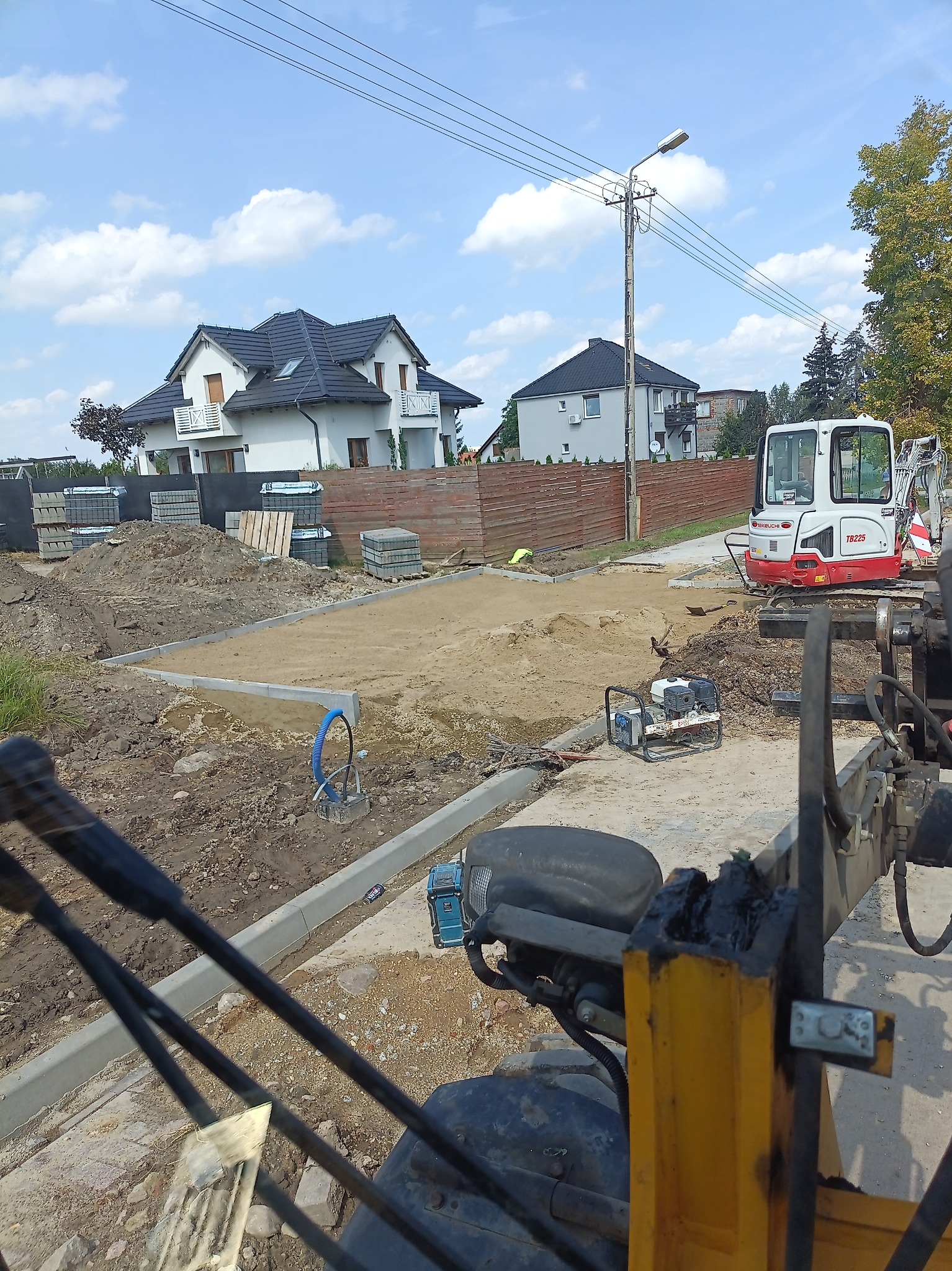 Przygotowany teren pod nawierzchnię z widocznymi krawężnikami, ubitym piaskiem, stertą kostki, mini koparką Takeuchi TB225 i maszyną budowlaną z żółtymi elementami w pierwszym planie, na tle domów...