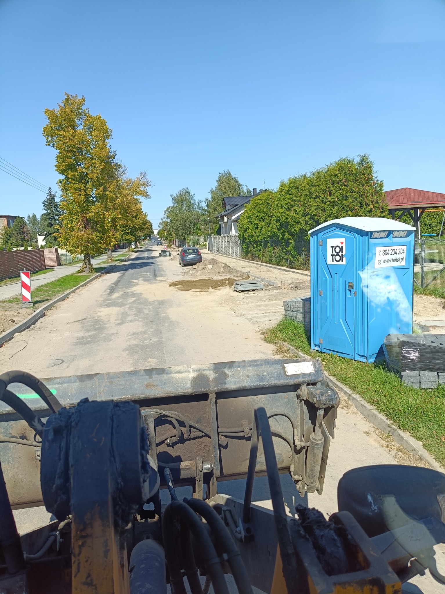 Przygotowanie podłoża pod kostkę brukową na ulicy w słoneczny dzień, widok z kabiny maszyny budowlanej, z przenośną toaletą Toi Toi w tle.