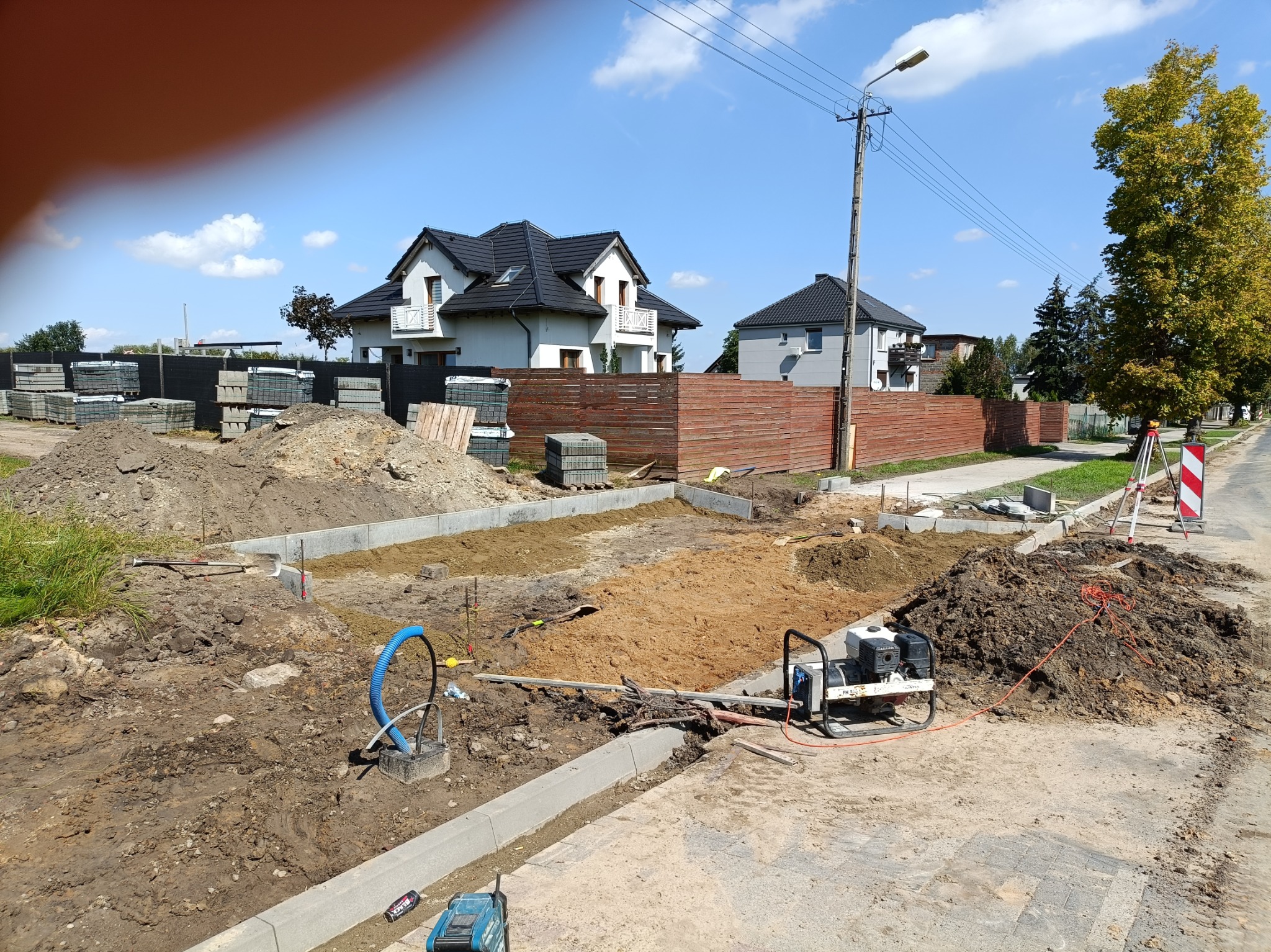 Przygotowanie terenu pod budowę chodnika lub drogi, widoczne krawężniki, hałdy ziemi, ubijarka, niebieski przewód, w tle domy jednorodzinne i stosy kostki brukowej.
