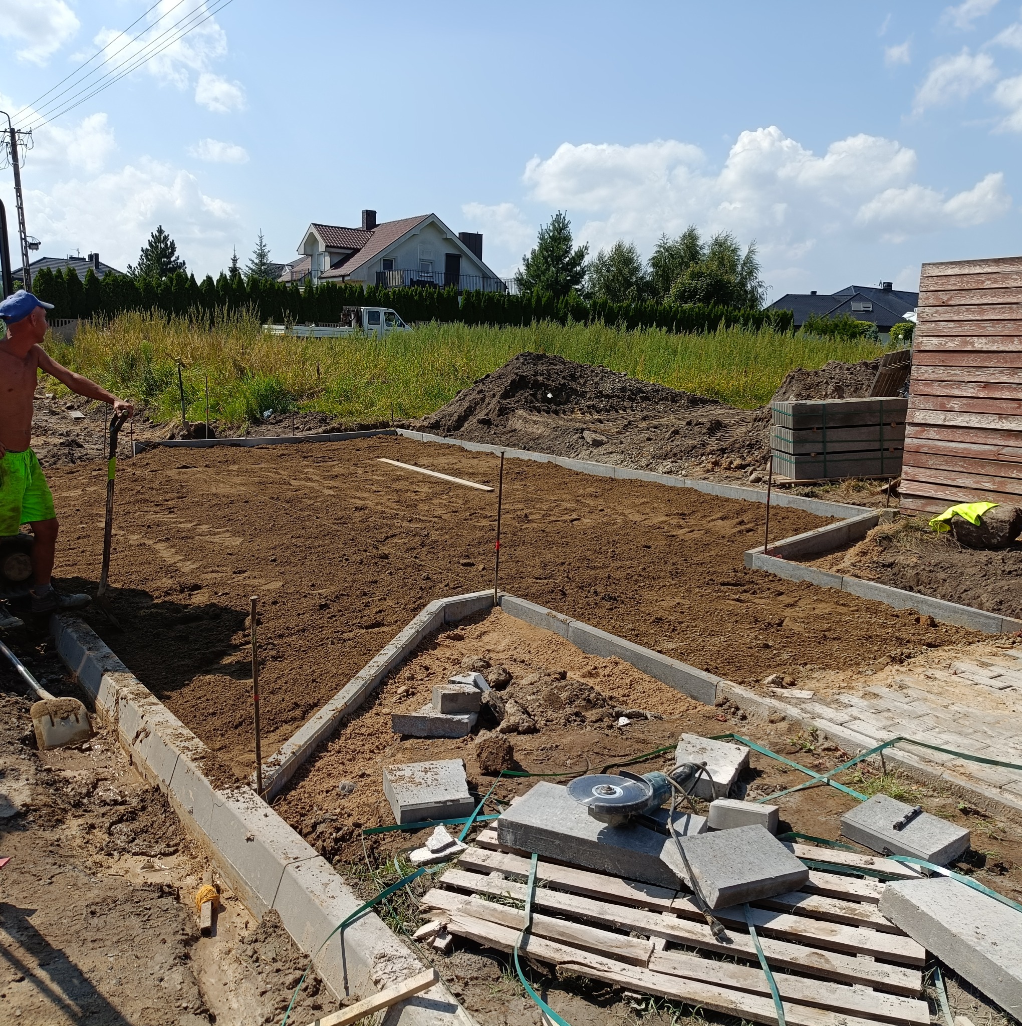 Przygotowanie terenu pod nawierzchnię z obrzeżami, widoczny robotnik z łopatą, wyrównany piasek, paleta z elementami nawierzchni i narzędzie do cięcia.