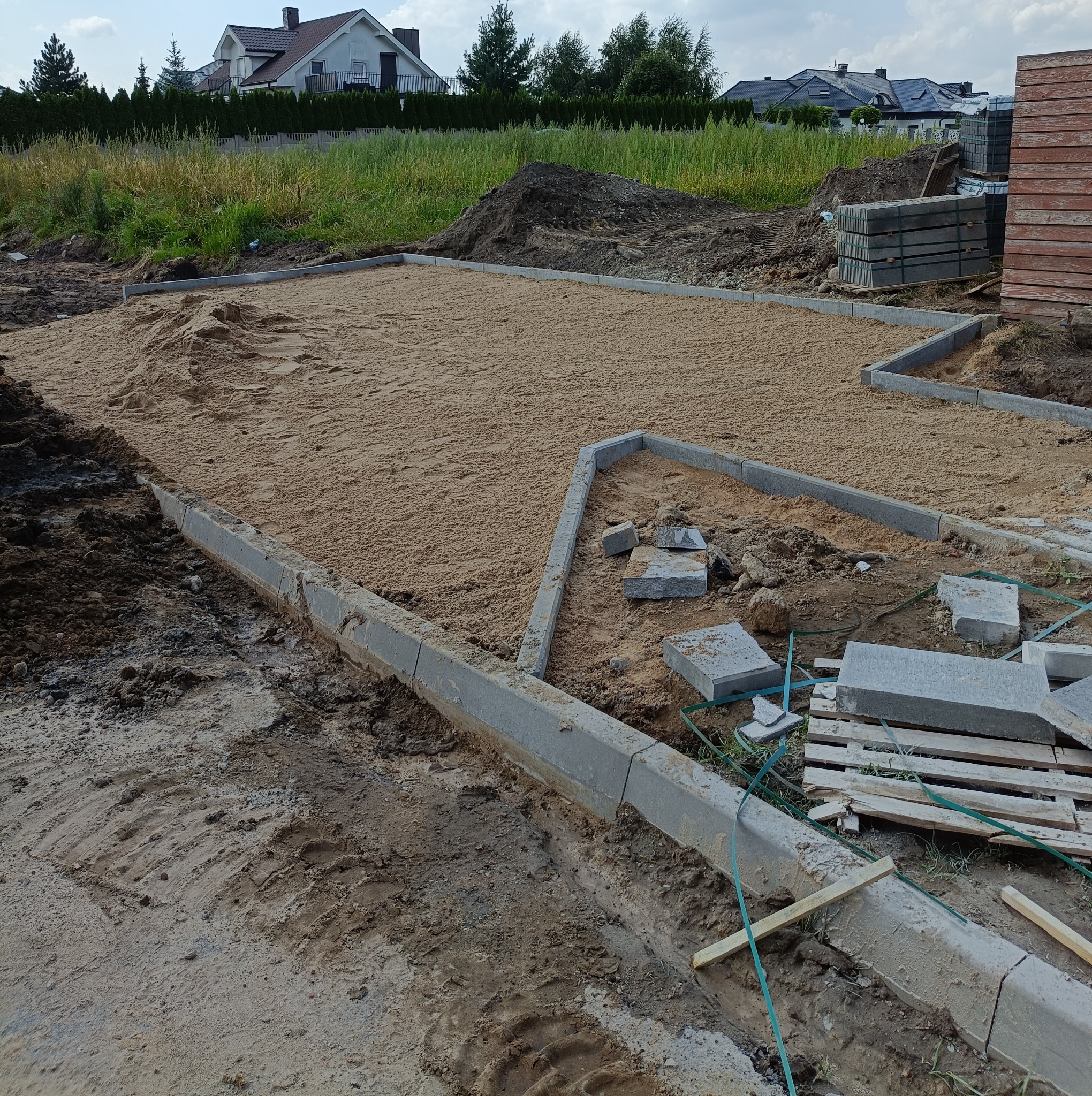 Przygotowanie podłoża pod nawierzchnię: wyrównany piasek ograniczony betonowymi obrzeżami, paleta z kostką w tle, widoczne domy w oddali.