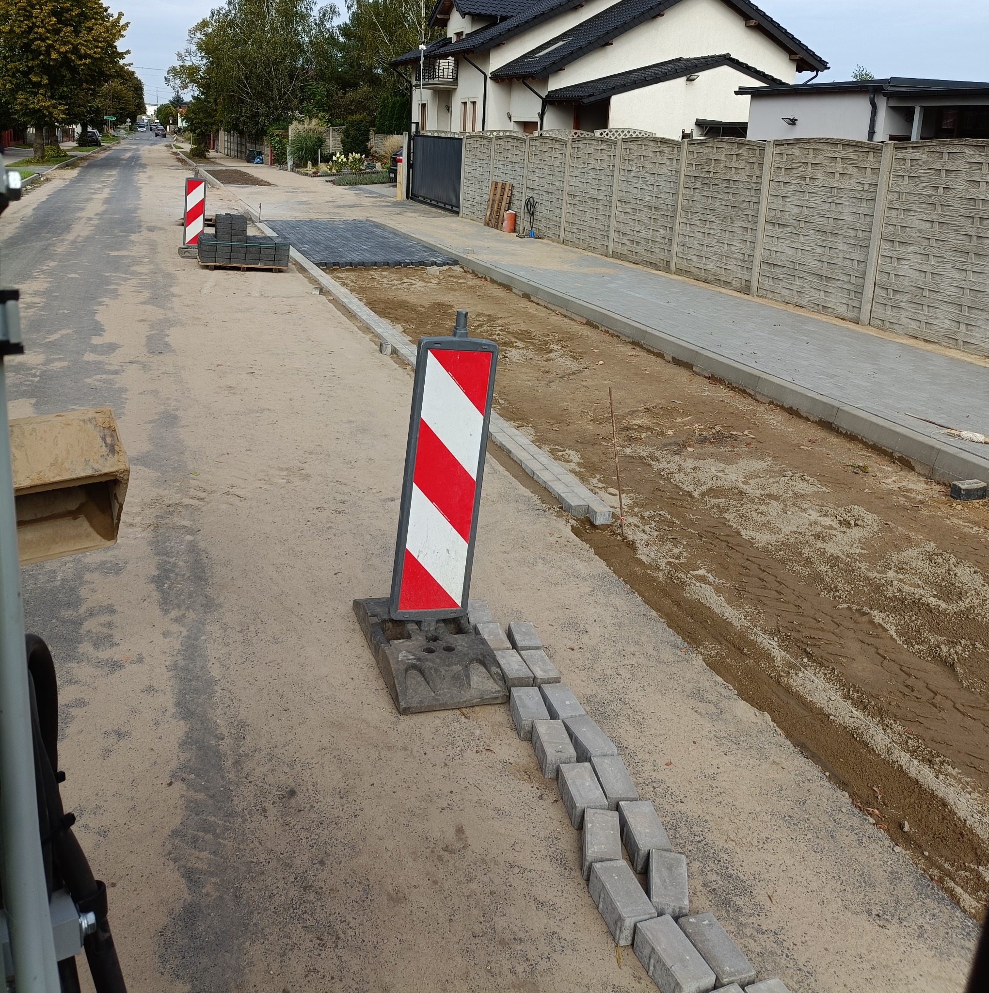 Ulica w trakcie remontu, widoczne przygotowane podłoże, ułożone obrzeża chodnika, paleta z kostką brukową i znaki ostrzegawcze, perspektywa z kabiny maszyny budowlanej.