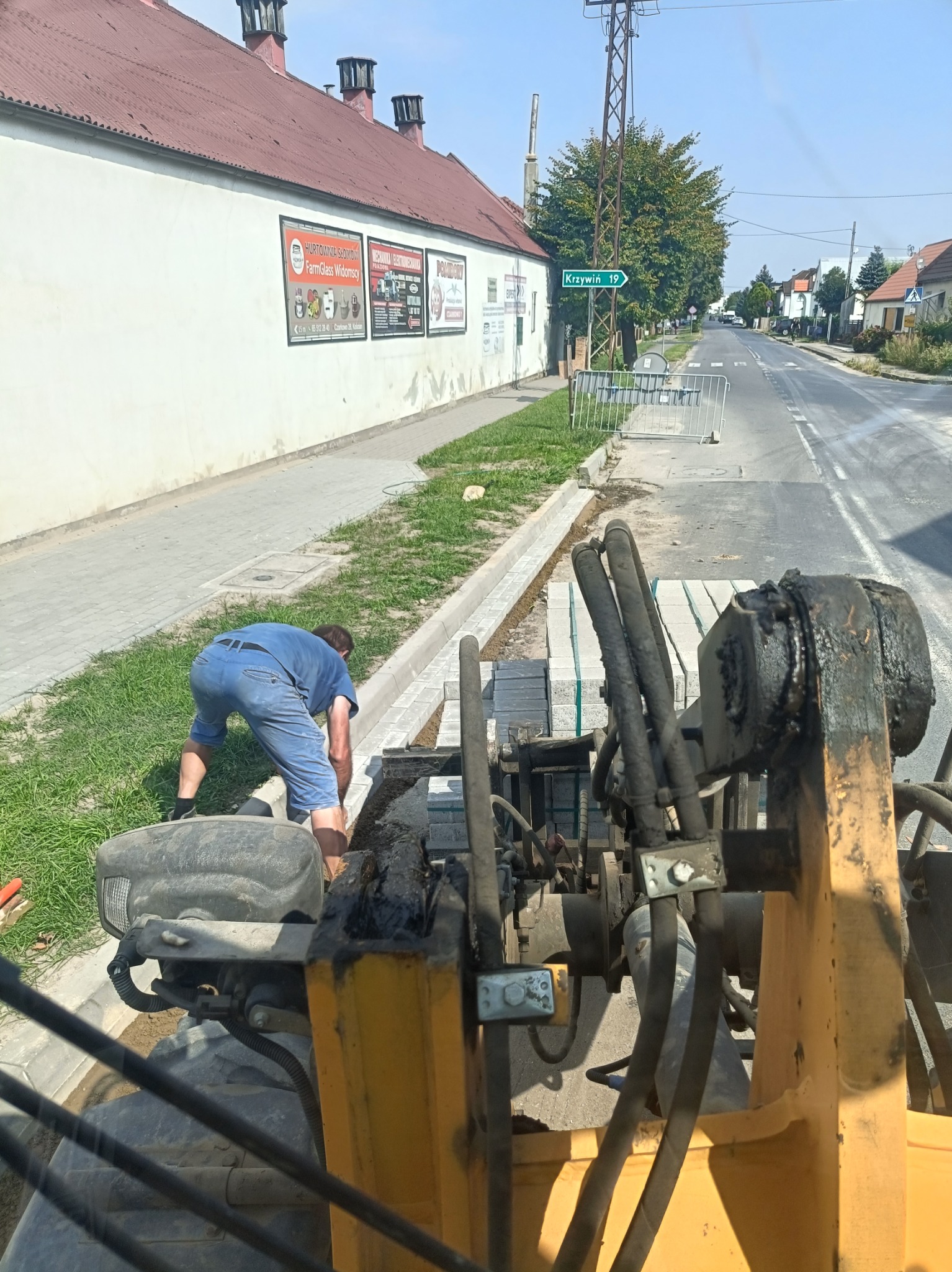 Pracownik w niebieskim kombinezonie układa szare kostki przy krawężniku, widok z perspektywy operatora maszyny budowlanej, w tle ulica z tablicą kierunkową Krzywiń.