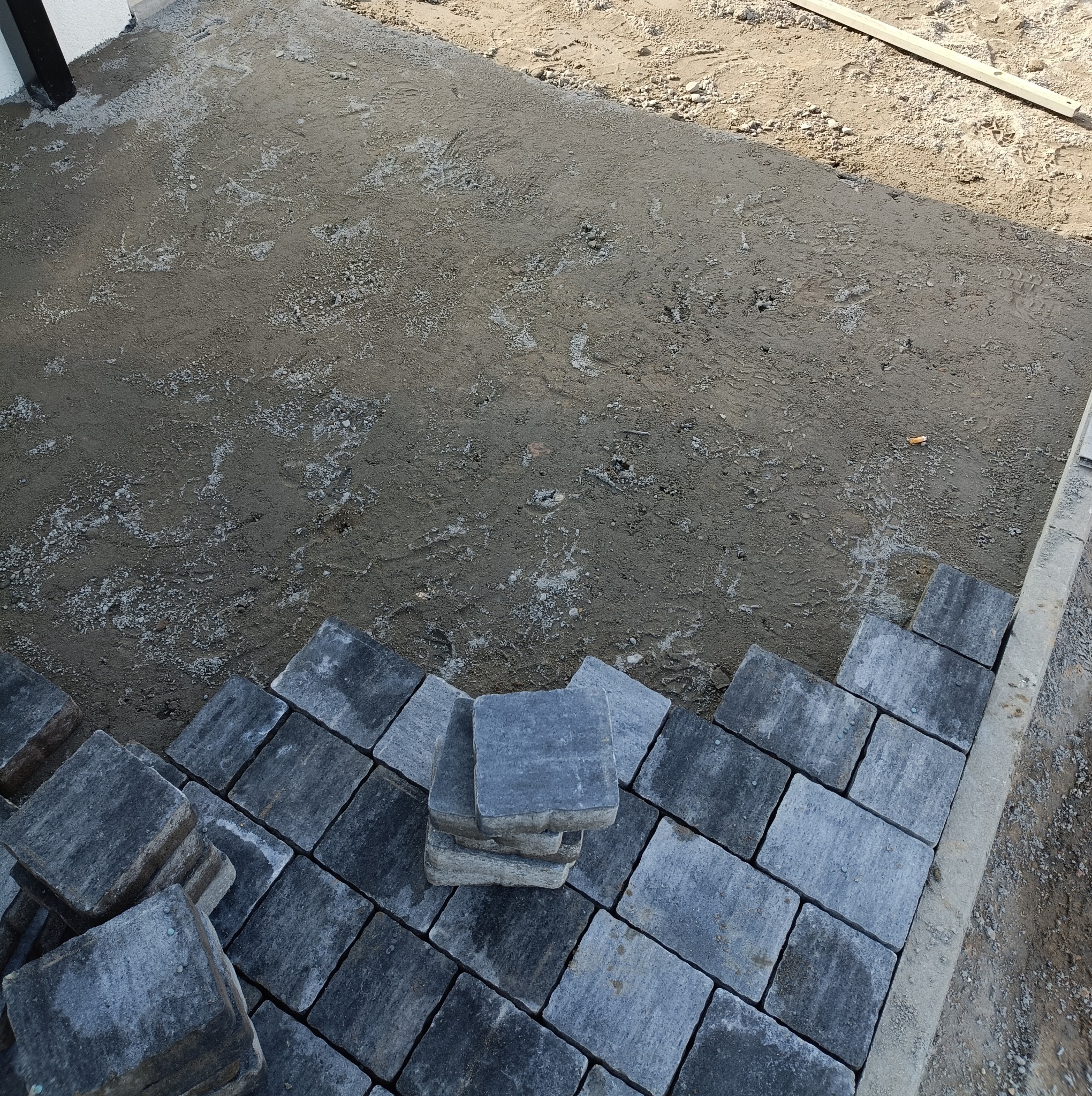 Układanie szarej kostki brukowej na podsypce piaskowej, widoczne ułożone fragmenty i stos kostki do dalszej pracy.