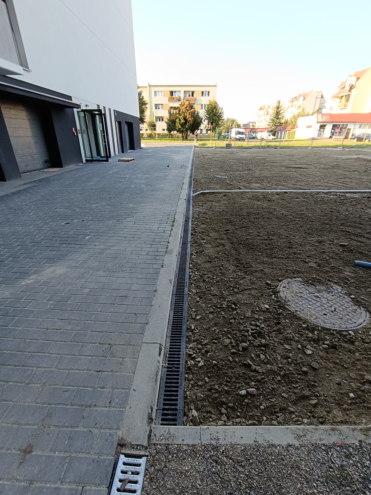 Szara kostka brukowa ułożona przy budynku, obok rów odwadniający i przygotowany teren pod przyszłą nawierzchnię z widocznym włazem kanalizacyjnym.