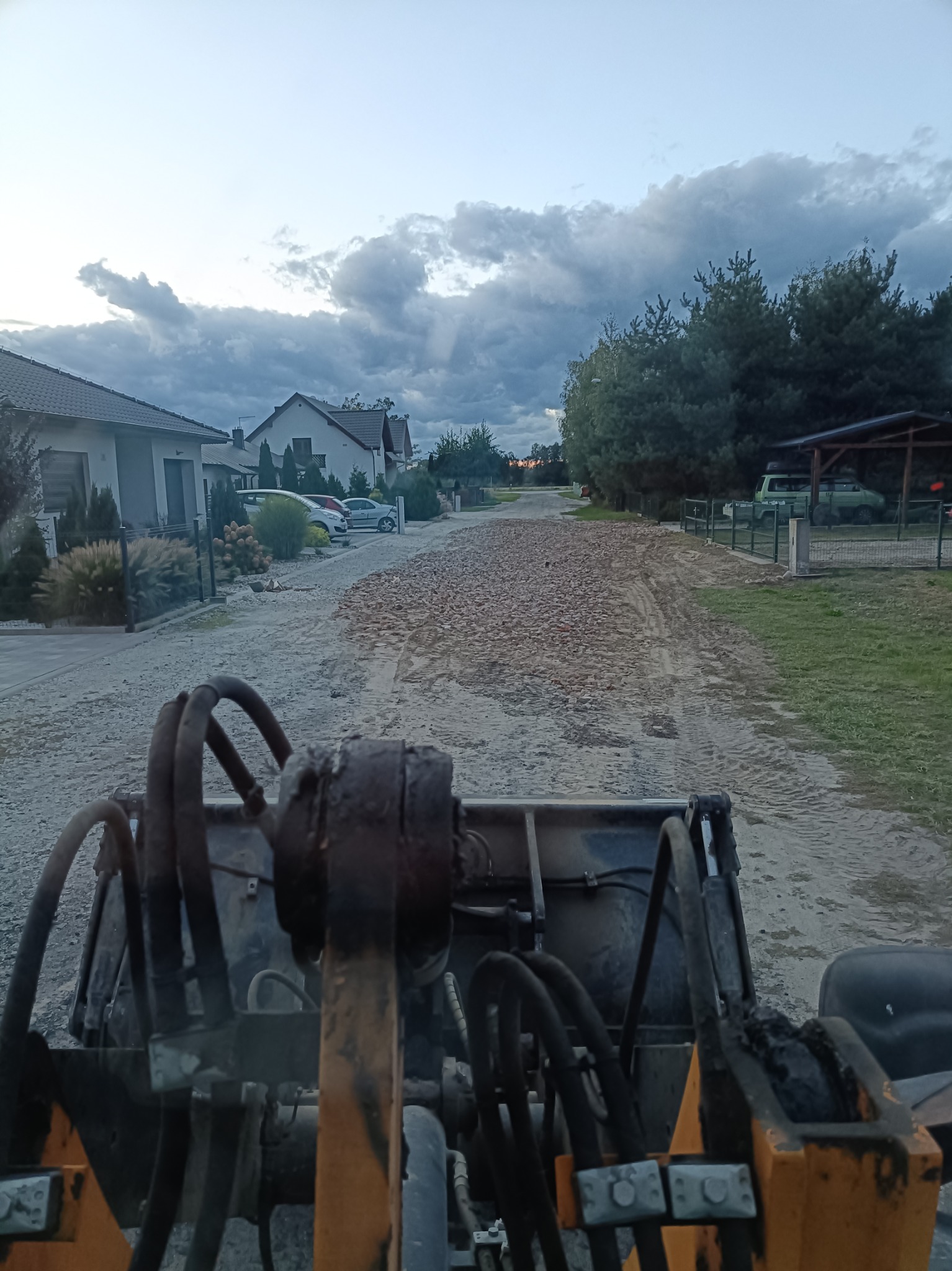 Widok z kabiny maszyny budowlanej na drogę gruntową z wysypanym gruzem, przygotowywaną do utwardzenia na tle domów jednorodzinnych i pochmurnego nieba.