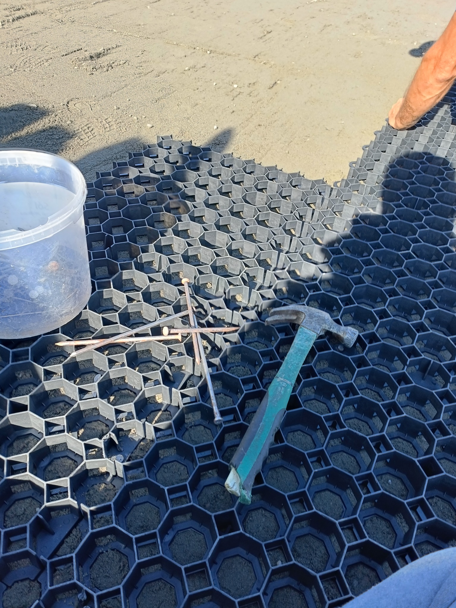 Układanie czarnej, plastikowej geokraty na piasku, z widocznym fragmentem ułożonej już powierzchni, obok wiadro z kołkami i młotek z zieloną rączką.