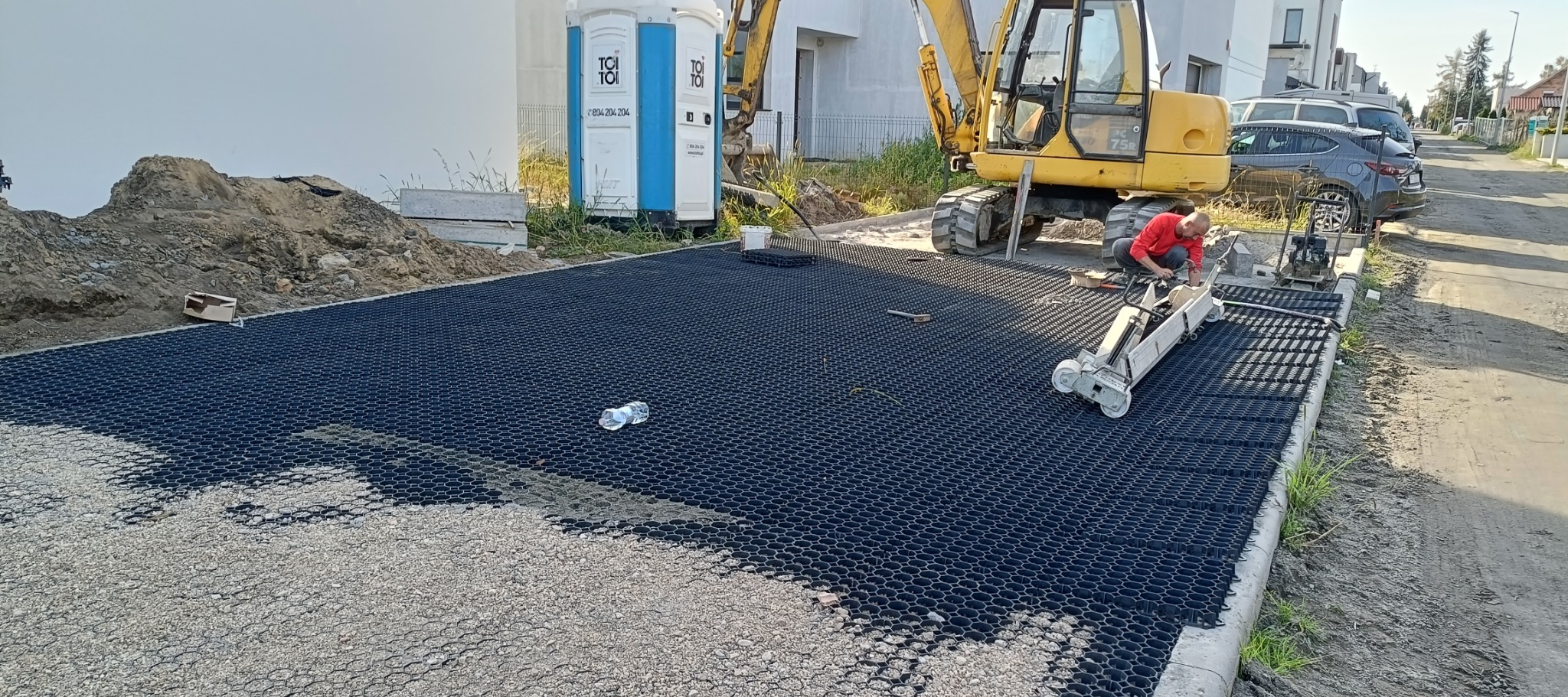 Pracownik w czerwonej koszulce układa czarne, ażurowe panele na podsypce z kruszywa, przy użyciu białej maszyny, obok stoi żółta koparka, w tle przenośna toaleta i budynki.