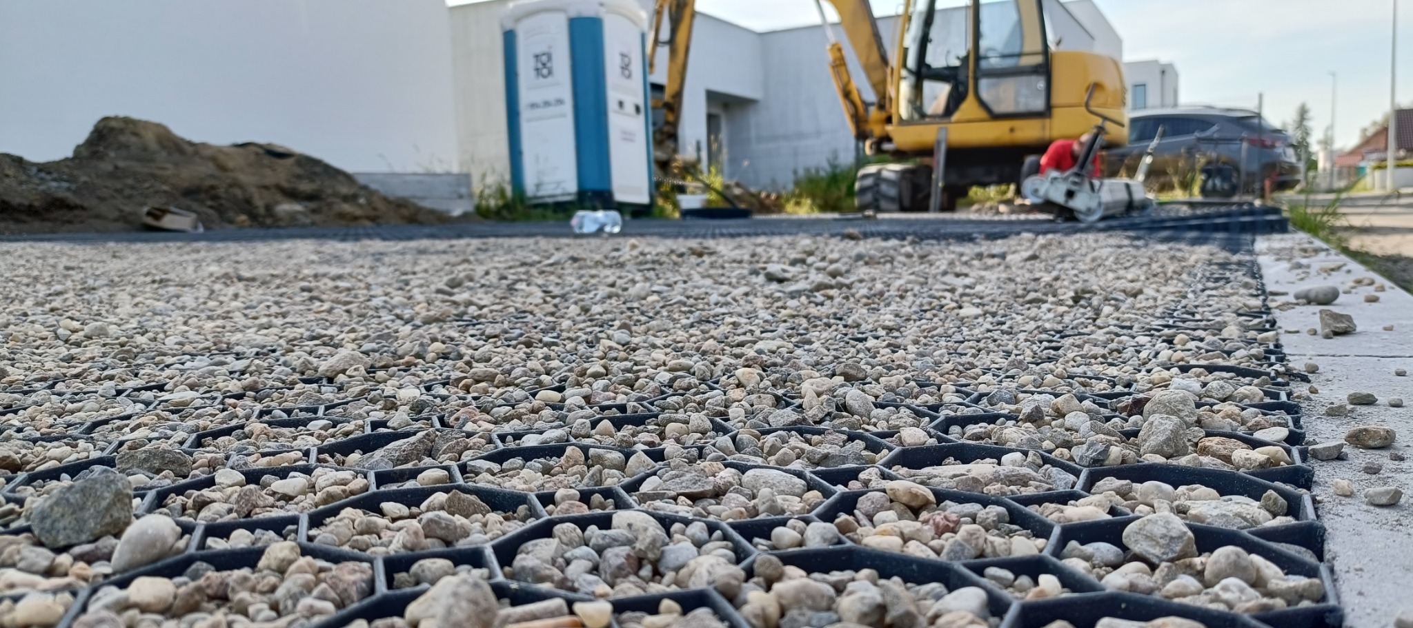 Geokrata wypełniona drobnym kruszywem, widok z bliska na strukturę, w tle koparka i przenośna toaleta, teren budowy.
