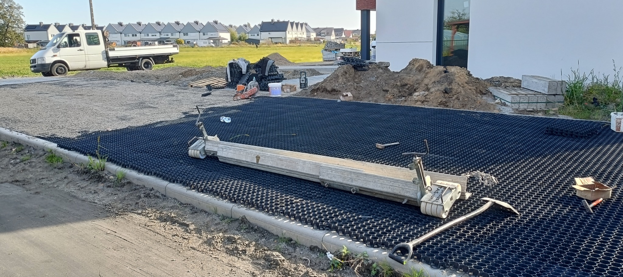 Układanie czarnej, plastikowej geokratki na przygotowanym podłożu, widoczne narzędzia i sprzęt budowlany, w tle domy i zaparkowany biały samochód dostawczy.