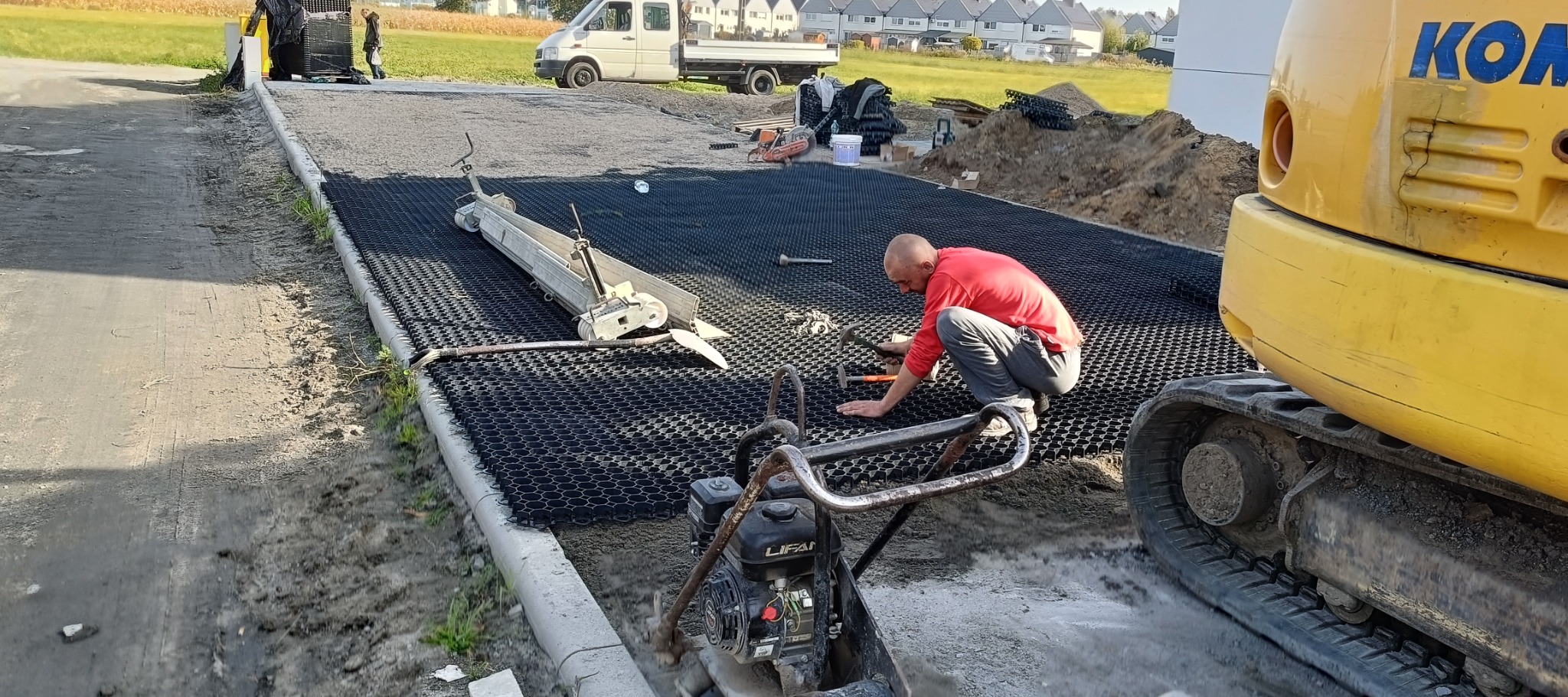 Pracownik w czerwonej koszulce wyrównuje podłoże pod przyszłą nawierzchnię z wykorzystaniem kratki trawnikowej, widoczna ubijarka, mini koparka Komatsu, listwa wyrównująca, w tle zabudowa...