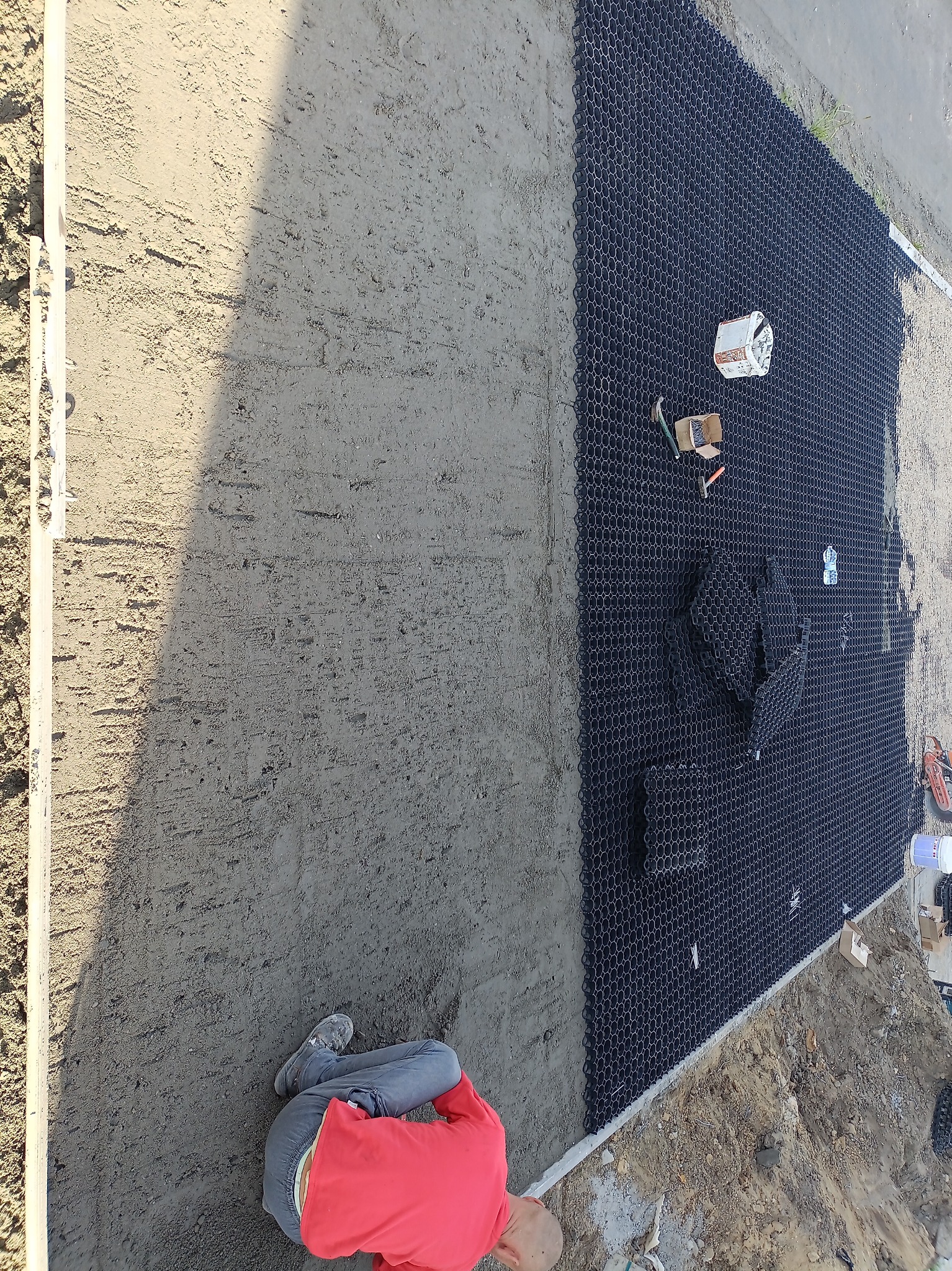 Montaż czarnej, plastikowej geokratki na przygotowanym podłożu. Widoczne narzędzia, częściowo rozłożone elementy i pracownik w czerwonej bluzie.