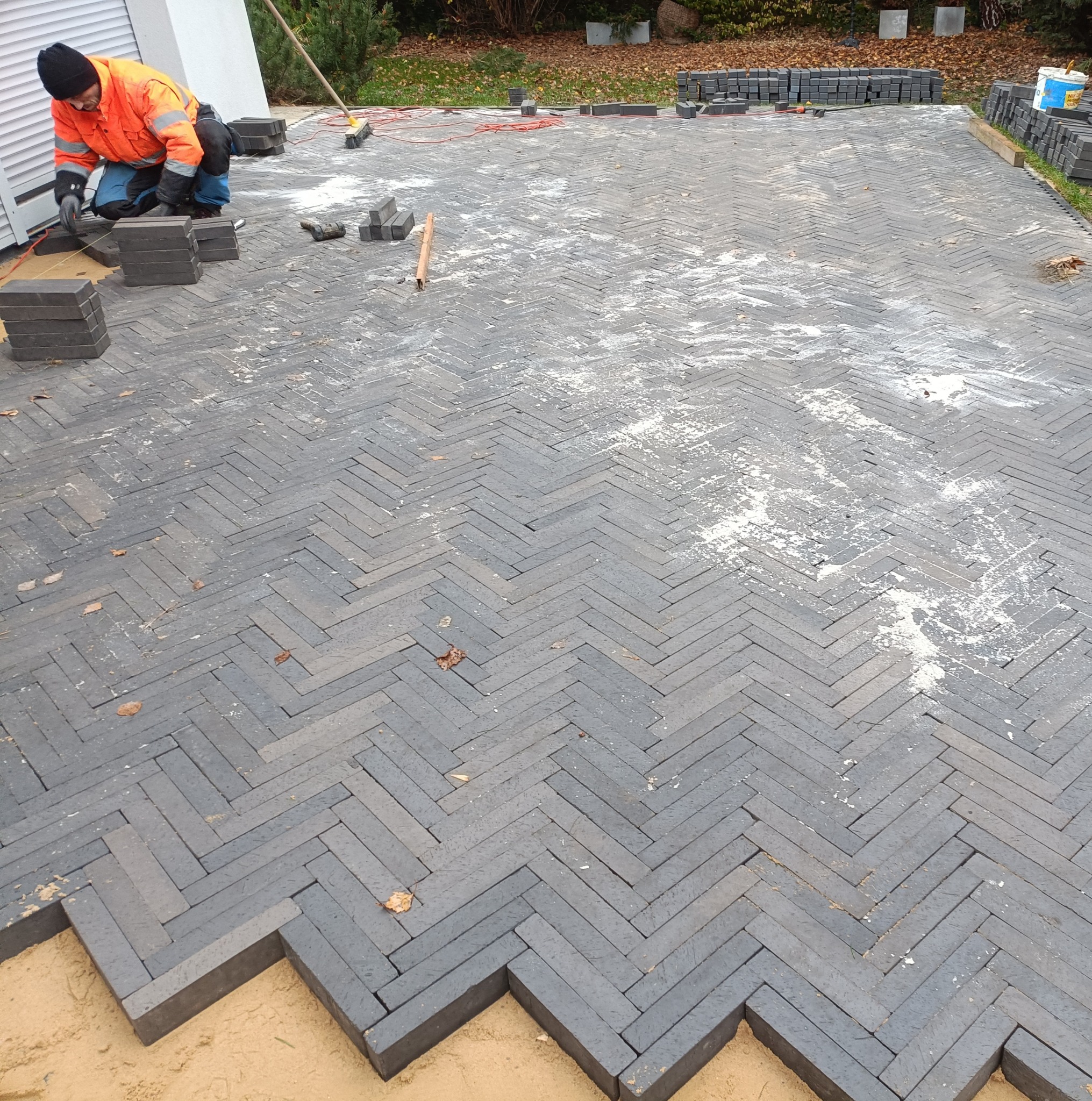 Układanie kostki brukowej w wzór jodełki z szarych elementów, widoczny pracownik w pomarańczowej kurtce i czapce, stosy kostki i narzędzia, podsypka piaskowa na obrzeżach.