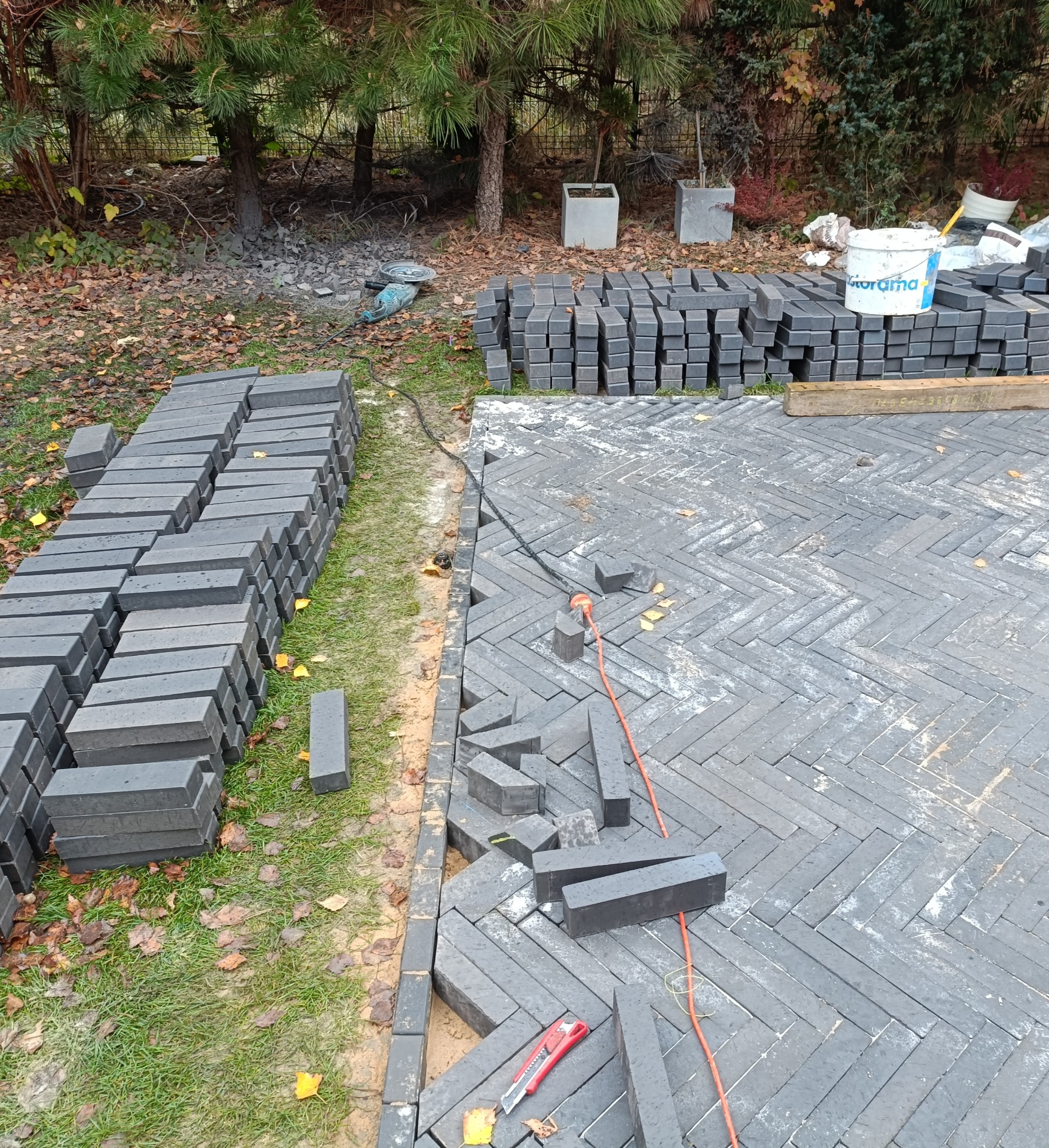 Ciemnoszara kostka ułożona we wzór jodełki, częściowo ukończona, z widocznymi zapasami kostki, narzędziami (nóż) i kablem zasilającym na trawie i ziemi. W tle drzewa i betonowe donice.