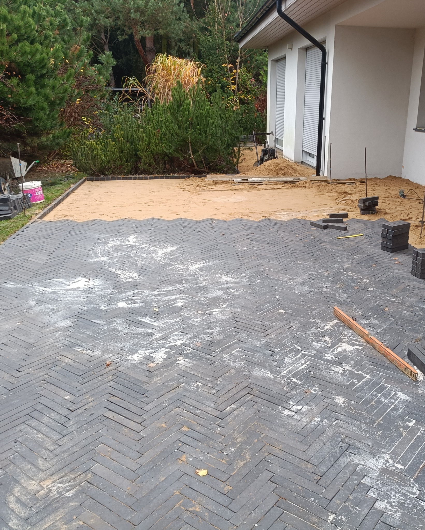 Częściowo ułożona nawierzchnia z ciemnoszarej kostki w jodełkę, podsypana piaskiem, z widocznym poziomem i stosem kostki, na tle domu z szarymi roletami i zielenią.