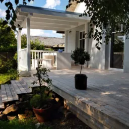 Drewniany taras z pergolą w jasnych odcieniach, z roślinami w donicach i widokiem na ogród.