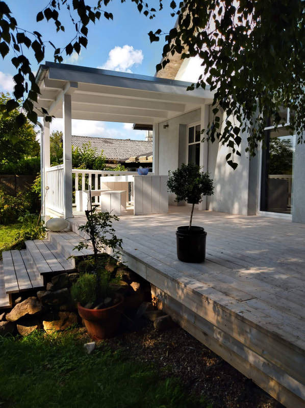 Drewniany taras z pergolą w jasnych odcieniach, z roślinami w donicach i widokiem na ogród.