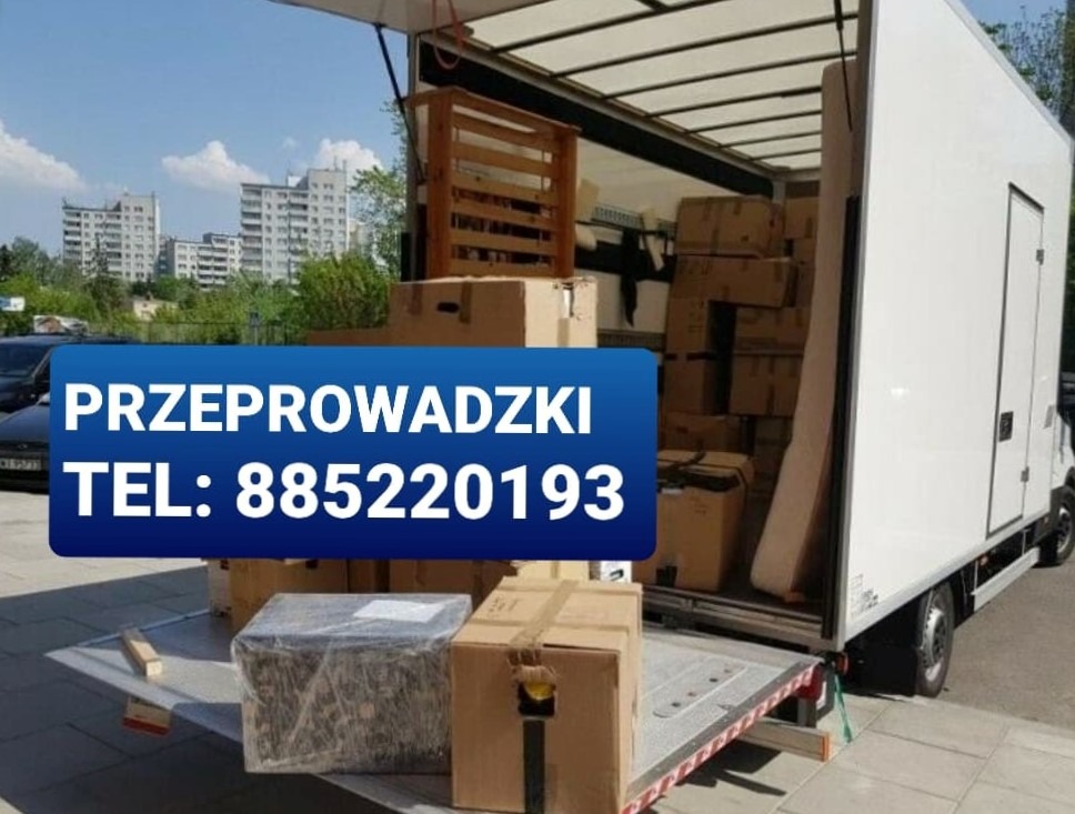 Otwarta paka białego samochodu dostawczego załadowana kartonami i drewnianą paletą, widok z zewnątrz na tle bloków mieszkalnych.