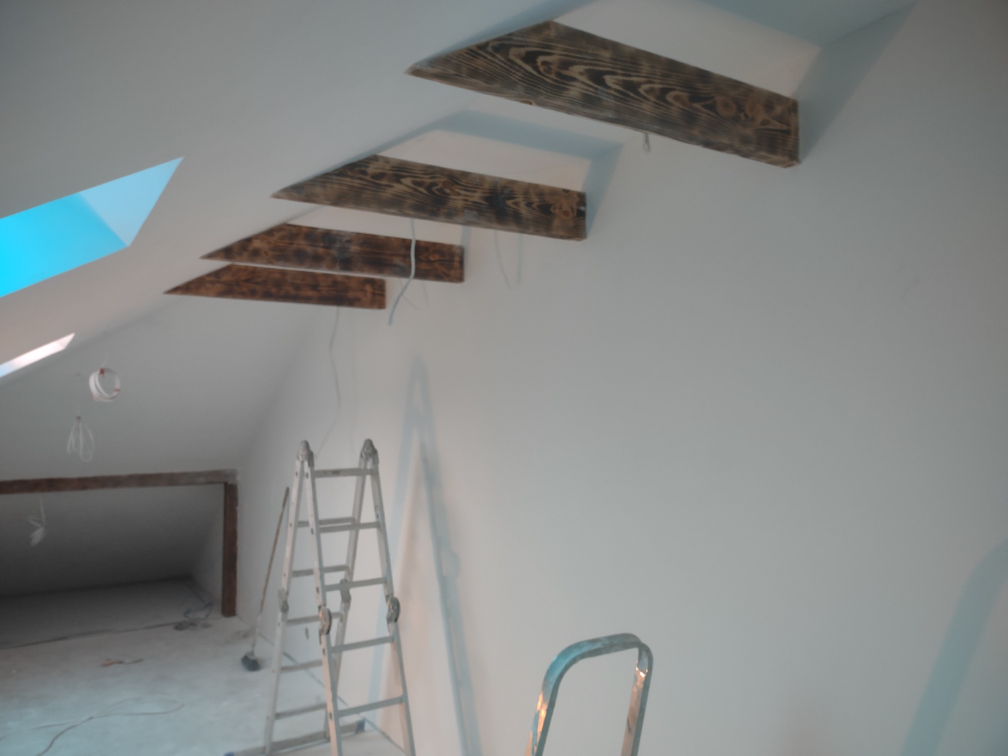 Poddasze w trakcie wykańczania: widoczne drewniane belki stropowe, ściany przygotowane do malowania, drabina aluminiowa, fragment okna dachowego.