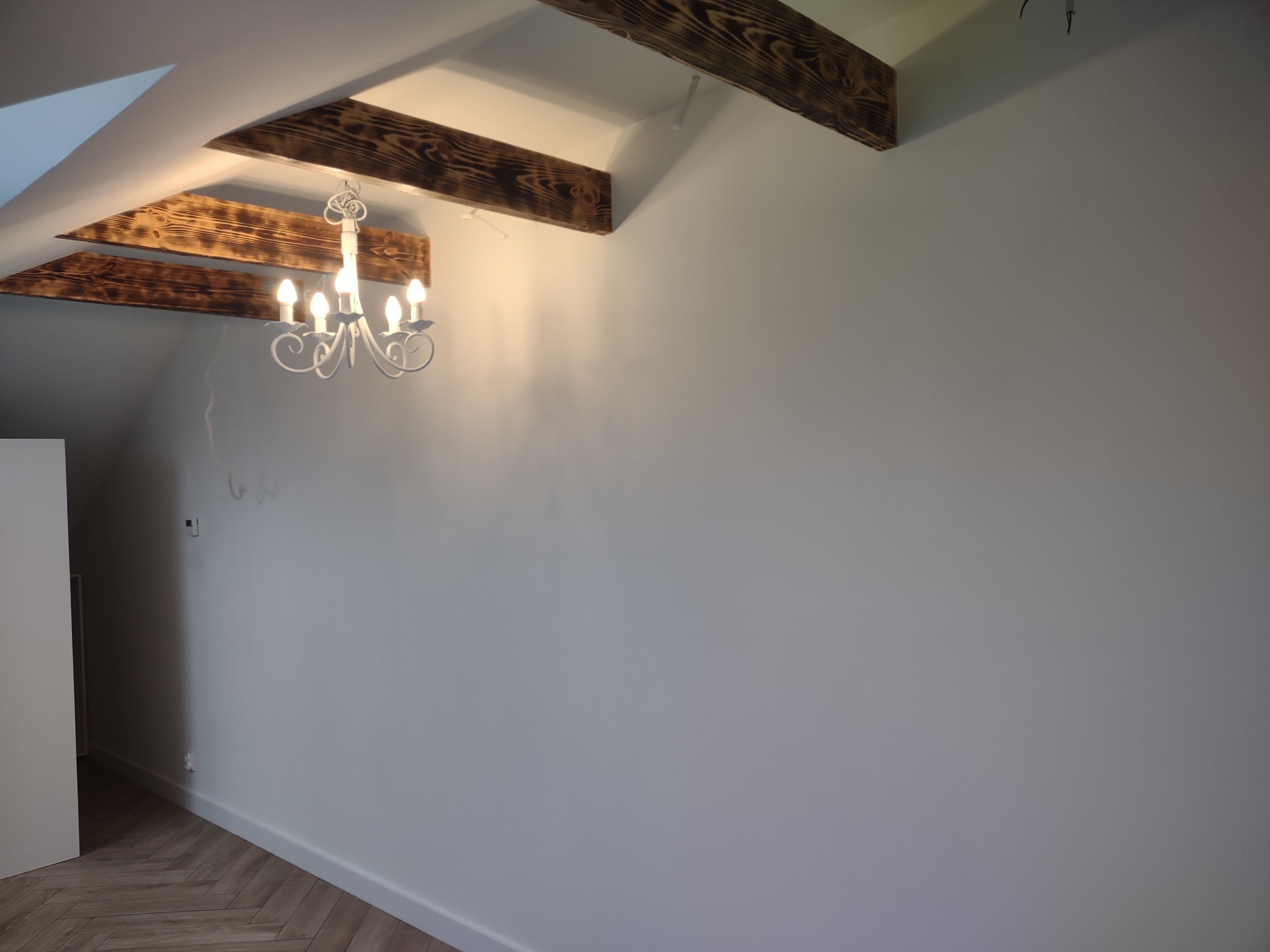Poddasze po remoncie z drewnianymi belkami stropowymi, białymi ścianami i parkietem w jodełkę; widoczny biały żyrandol.