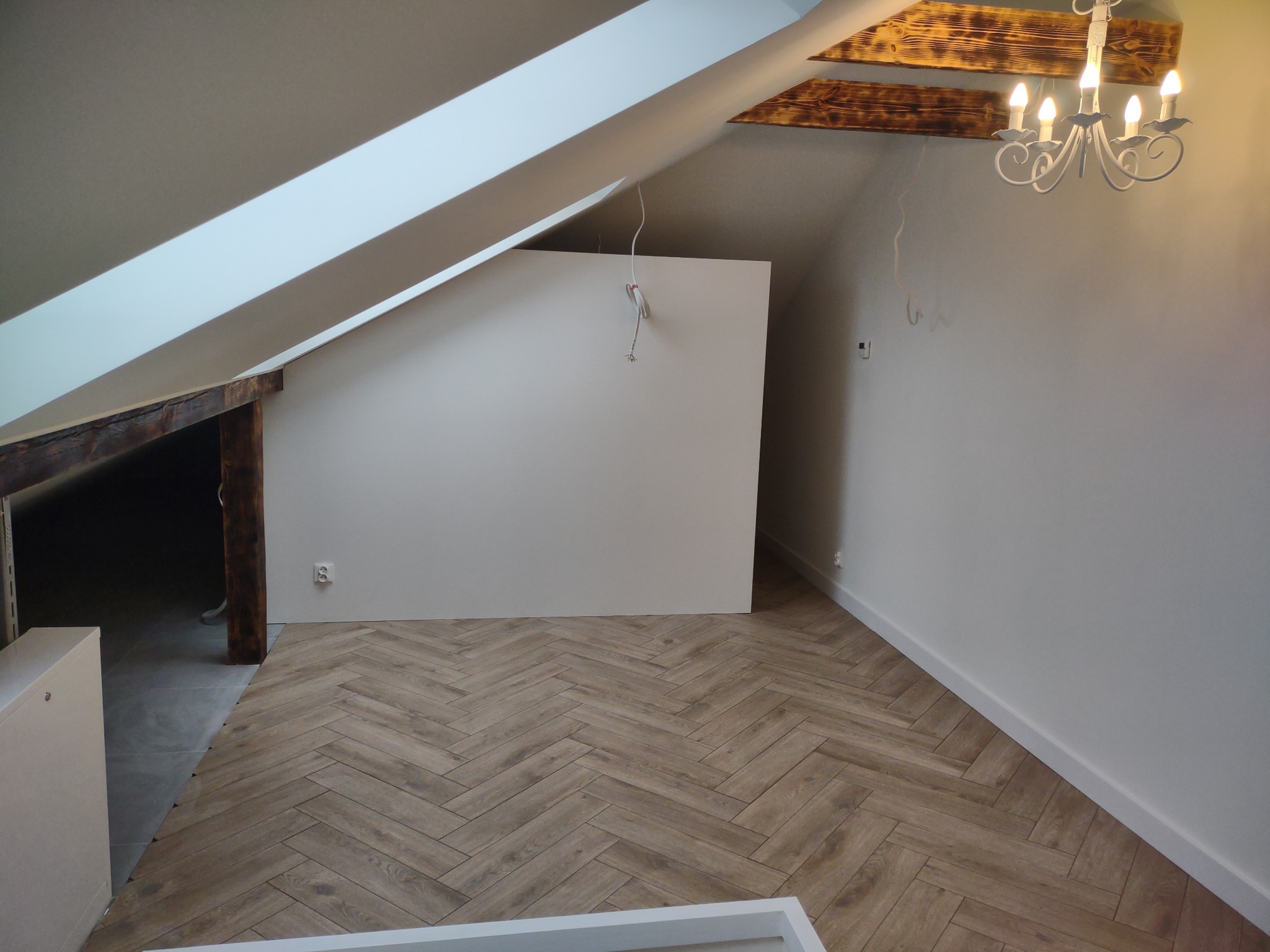 Poddasze po remoncie: drewniana podłoga ułożona w jodełkę, białe ściany, widoczne drewniane belki stropowe i biały żyrandol w stylu retro.