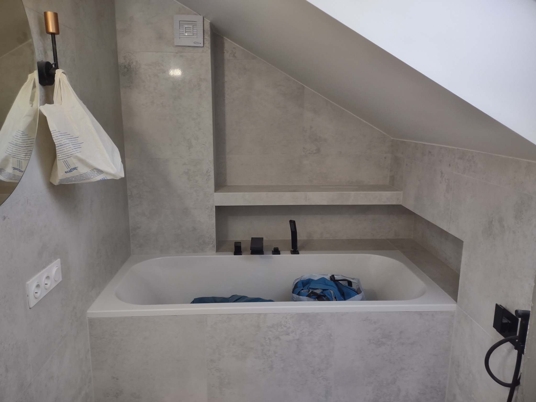 Wykończona łazienka na poddaszu z wanną obłożoną szarymi płytkami imitującymi beton, czarna bateria i deszczownica, półka w ścianie, biały skośny sufit, plastikowe torby wiszące na haku.