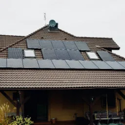 Brązowy dom z ciemnymi panelami słonecznymi na dachu z brązowej dachówki, widoczne okna dachowe i antena satelitarna.