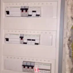 Rozdzielnica elektryczna natynkowa z bezpiecznikami, niektóre wyłączniki w pozycji włączonej, inne wyłączone, widoczna czerwona dioda sygnalizacyjna, numery obwodów nad bezpiecznikami.