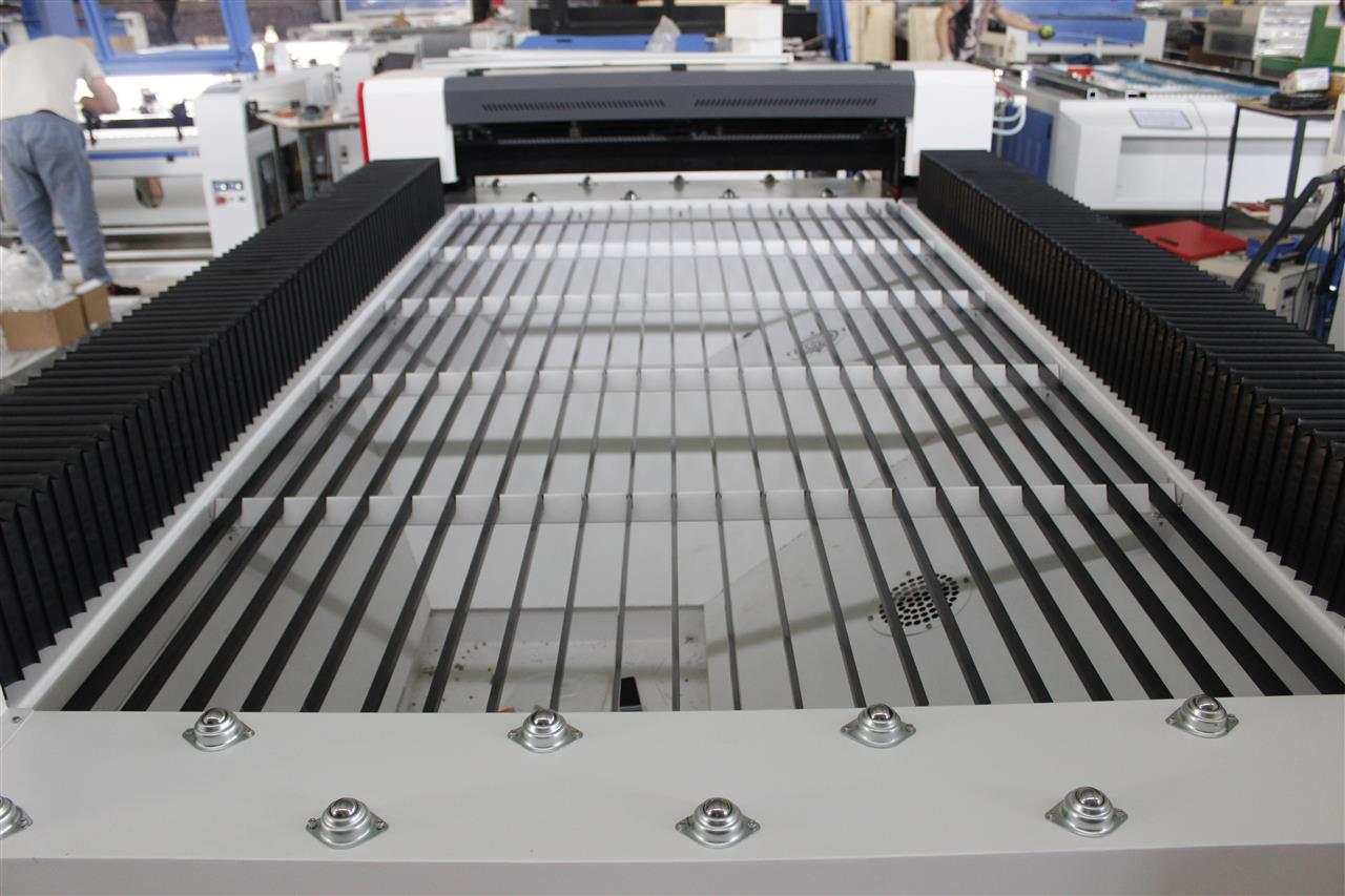 Szczegółowe ujęcie stołu roboczego maszyny do cięcia laserowego, z widocznymi czarnymi listwami i białymi przestrzeniami, otoczonego czarnymi osłonami harmonijkowymi, na tle hali produkcyjnej.