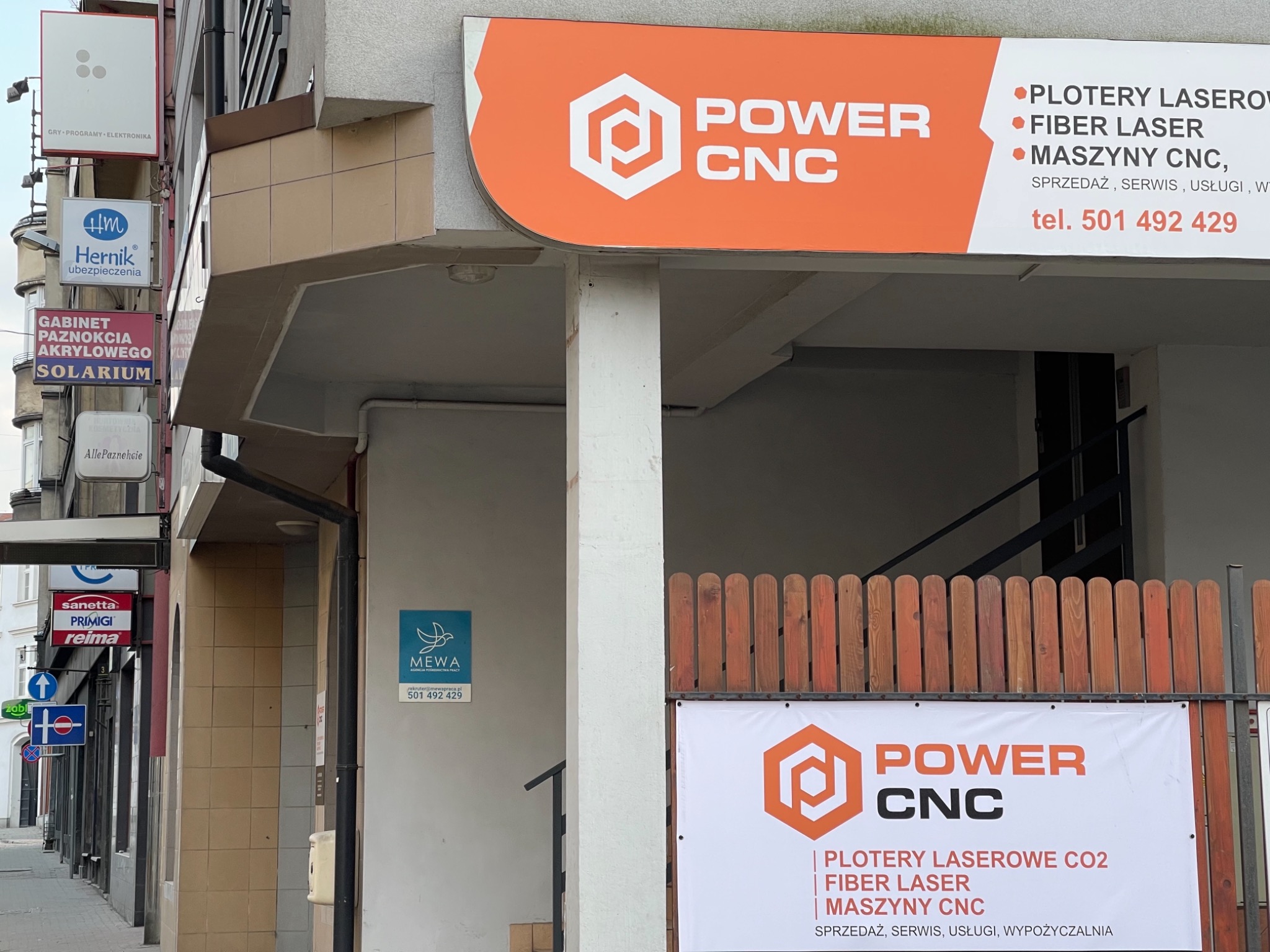 Zewnętrzny widok na budynek z szyldami firm, w tym szyld Power CNC oferującej plotery laserowe, fiber laser i maszyny CNC, widoczne także inne reklamy i znaki na ulicy.
