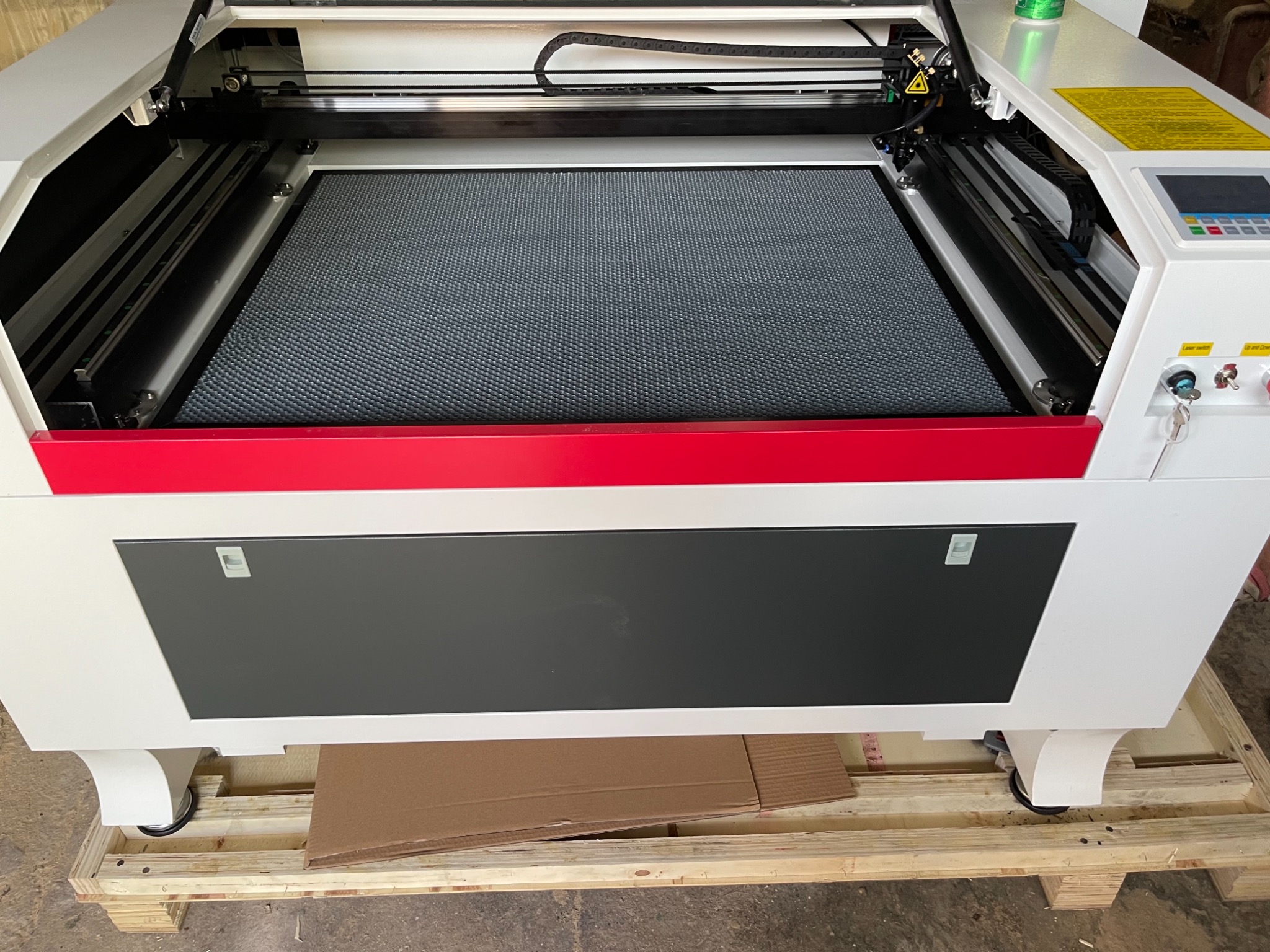 Wnętrze maszyny do cięcia laserowego z widocznym rusztem roboczym, elementami sterującymi i ostrzeżeniem laserowym, umieszczonej na drewnianej palecie transportowej.