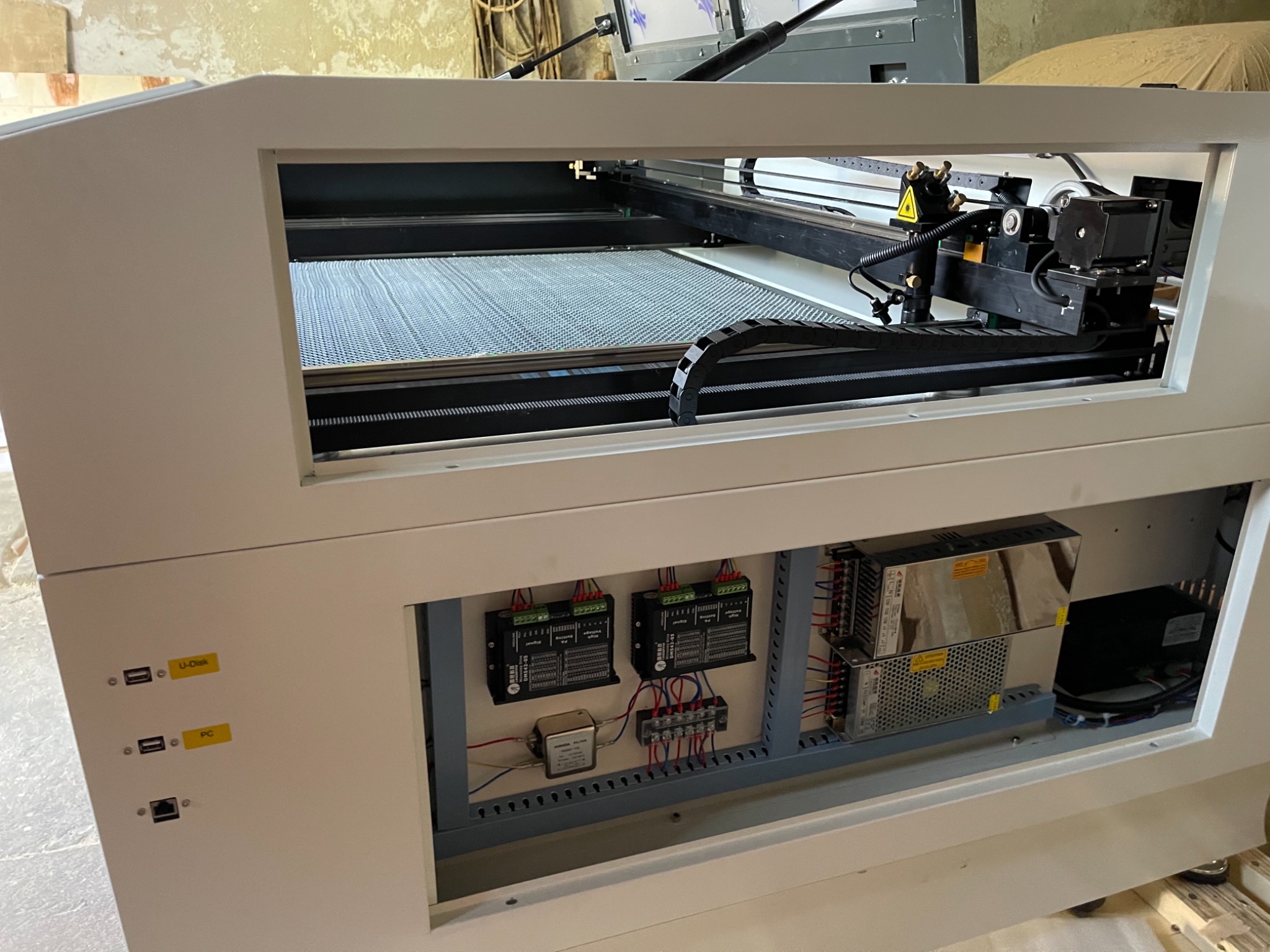 Wnętrze maszyny do cięcia laserowego z widocznym laserem, siatką roboczą i panelem elektrycznym zasilającym urządzenie.