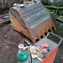 Ciężka, stalowa łyżka koparki po naprawie, z widocznymi świeżymi spawami, obok butla z gazem i wózek ze sprzętem ochronnym na zewnątrz w terenie.